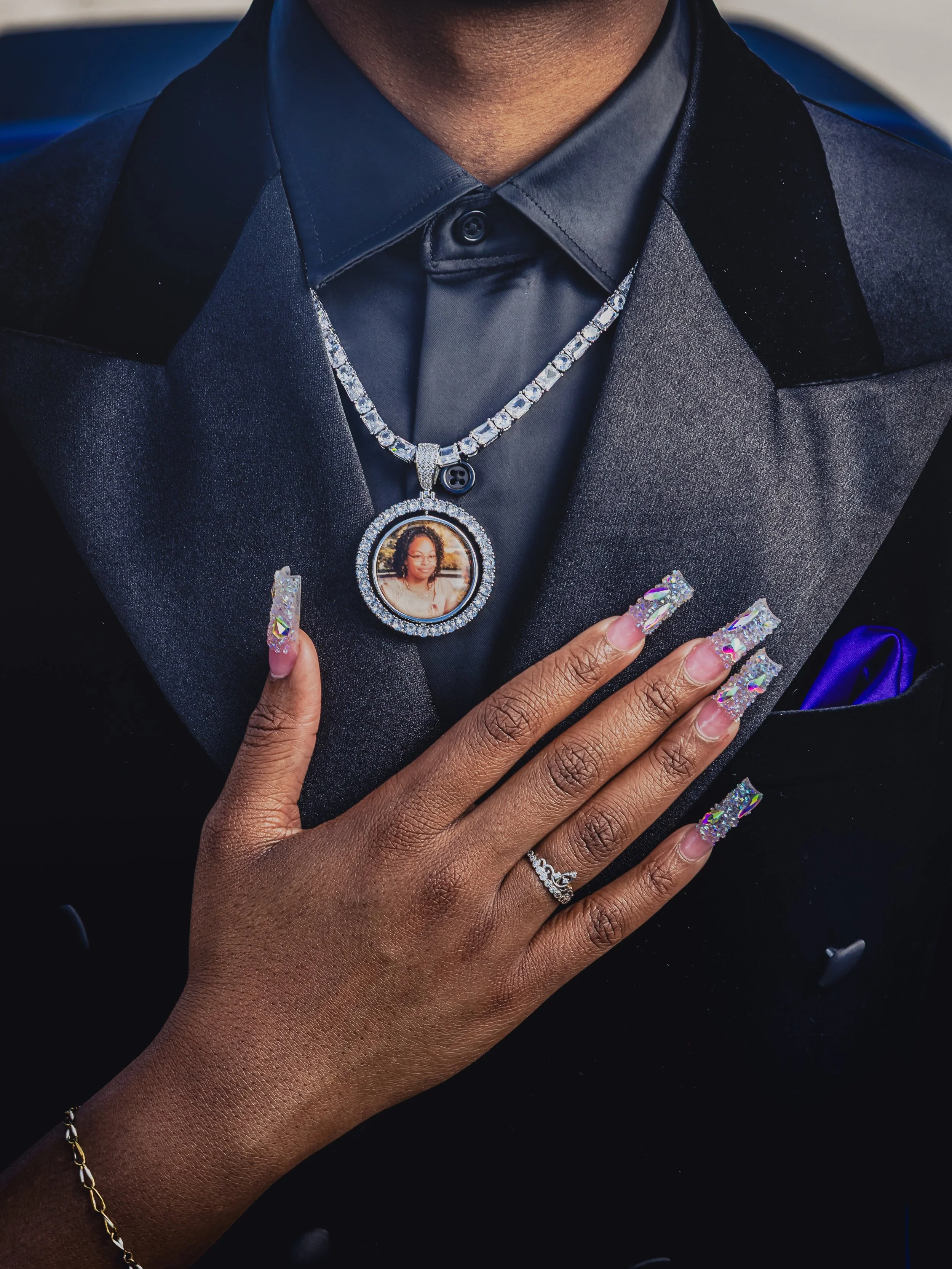 A woman with polished, glittery long nails and gold bracelet shows her hand on a man's chest. The man is wearing a dark gray suit with a black shirt and a blue pocket square. The man has a large diamond chain with a circular pendant featuring a woman