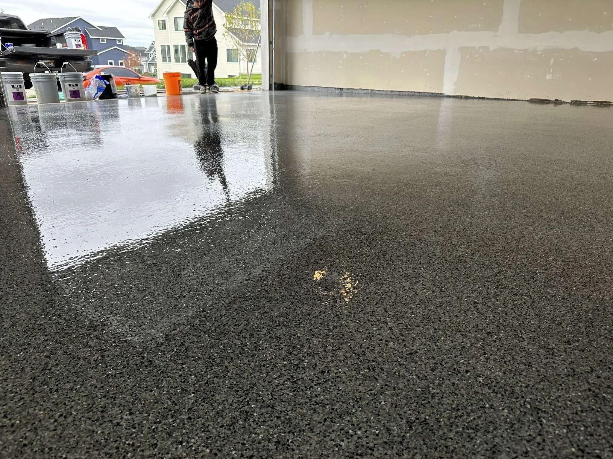 A freshly sealed concrete garage floor with a person walking in the background, several paint buckets and tools near the garage door, and an outdoor neighborhood scene visible through the open garage.