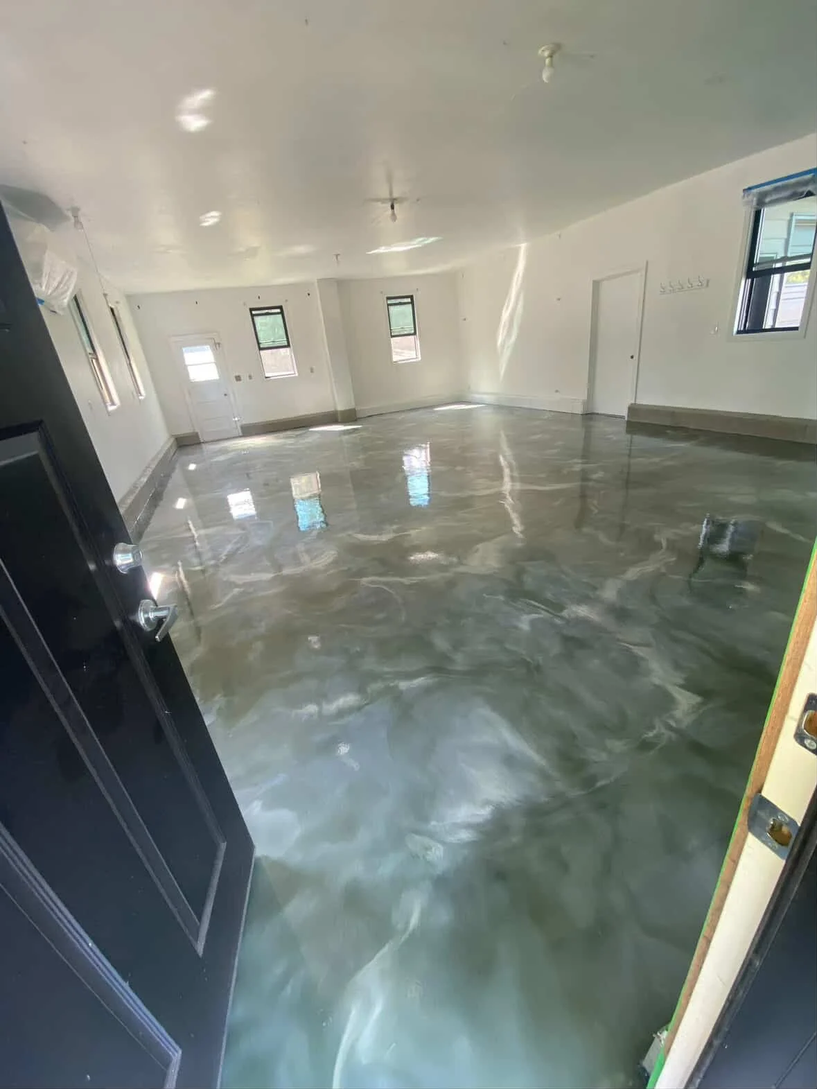 Empty room with shiny gray rice polished concrete floor, white walls, multiple windows with black frames, and a door at the back, appears to be under construction or renovation.