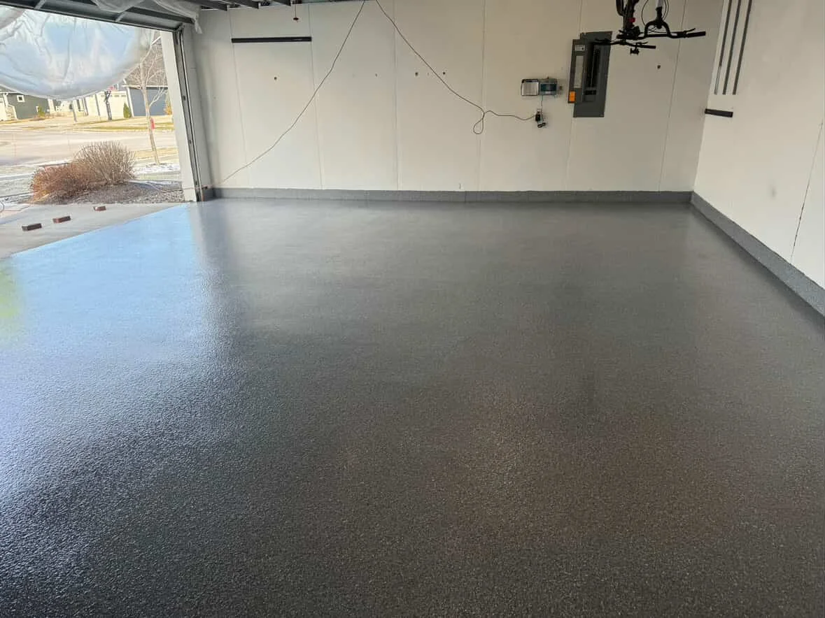 Empty garage with a freshly coated gray epoxy floor, white walls, and an open garage door showing a street and some bushes outside.