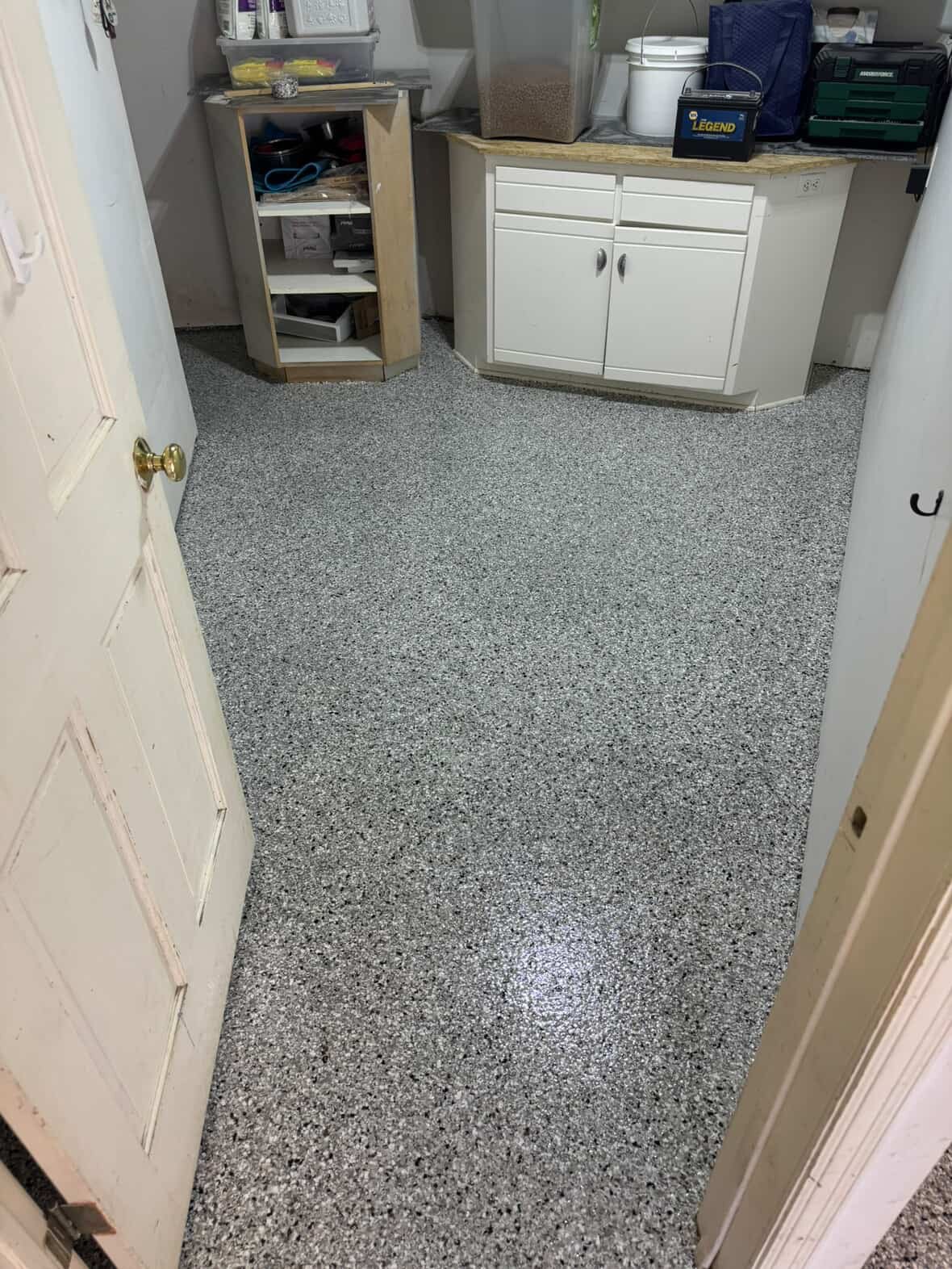 View of a small, cluttered utility room with speckled grey floor, white cabinets, and various storage containers and tools on counters and shelves.