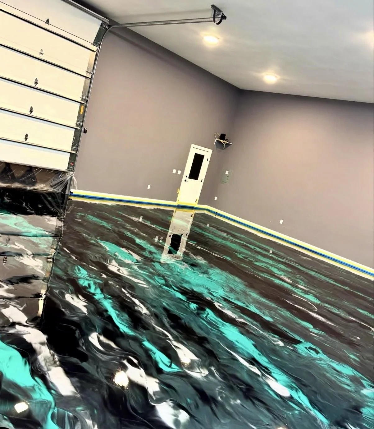 Interior of a garage with a shiny, custom epoxy floor with swirling black, gray, white, and teal colors, gray walls, a partially open garage door, ceiling lights, a white door with a window, a small wall-mounted shelf, and visible yellow and blue tap