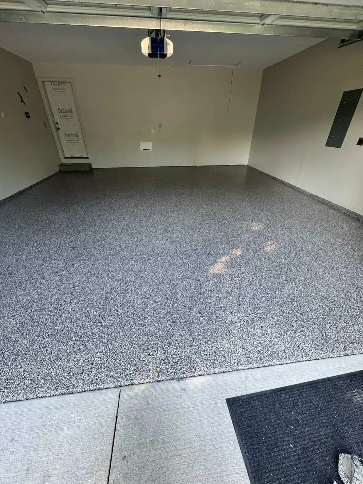 Empty garage with gray speckled epoxy floor, white walls, and a closed door. Garage door opener hanging from the ceiling and some light reflections on the floor.