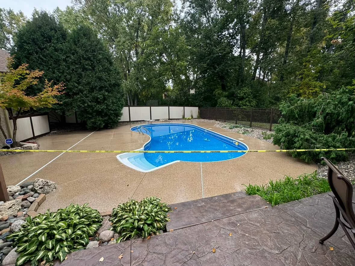 Empty backyard swimming pool surrounded by a concrete deck, with a caution tape across the middle. The pool is blue with a curved shape, situated in a landscaped yard with plants, trees, and a fence.