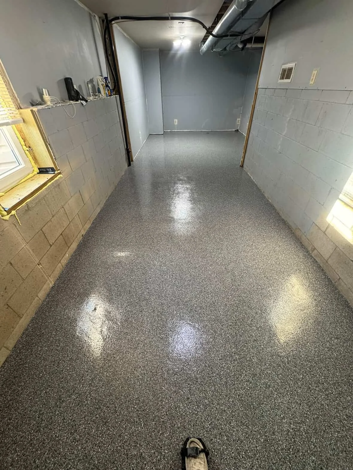 Empty unfinished basement with polished epoxy flooring, cinder block walls, exposed pipes, a window, and a single shoe in the foreground.