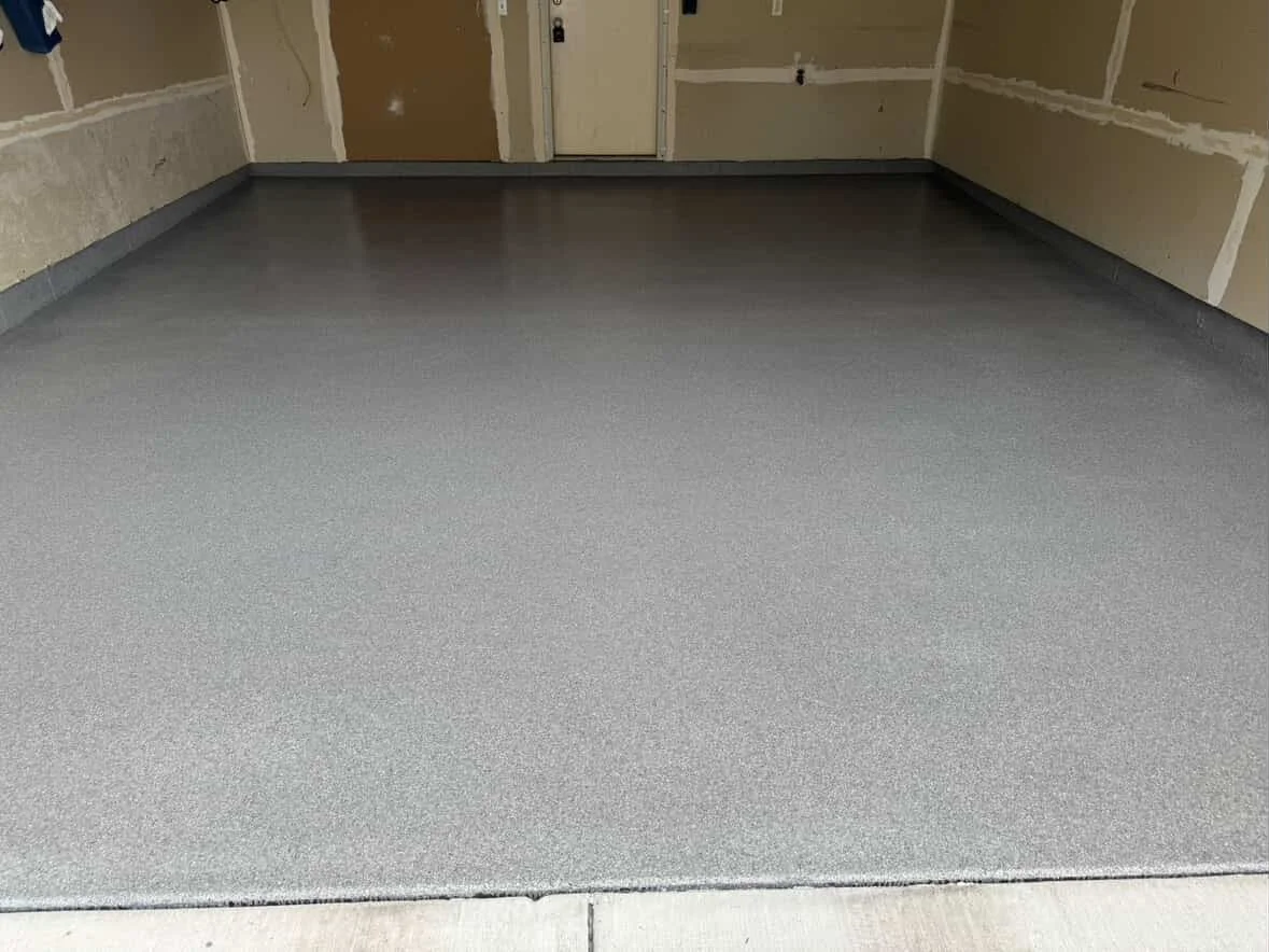 Interior of an unfinished garage with freshly coated gray concrete floor, unfinished walls, and a door in the background.