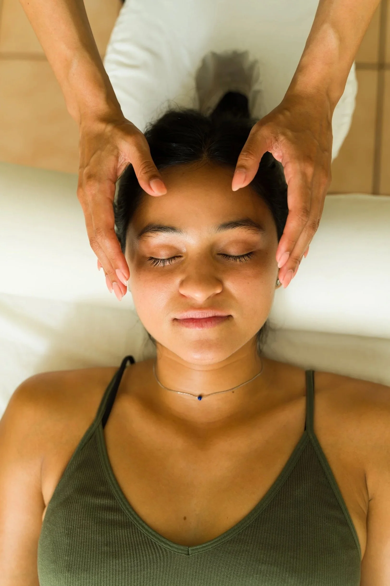 A woman lying on her back with eyes closed, receiving a massage on her forehead and temples.