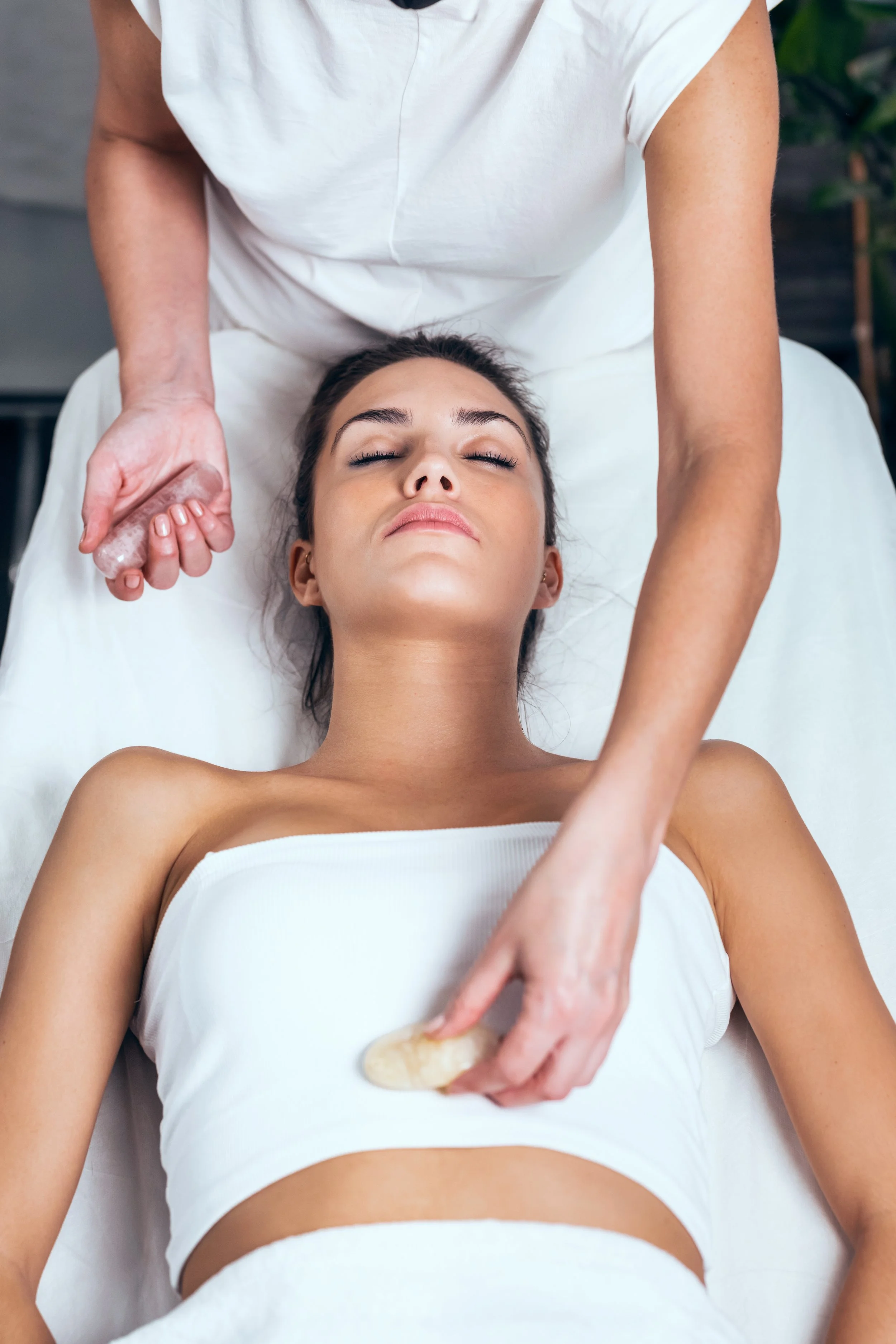 A woman lying down with her eyes closed, receiving a facial massage from a therapist who is using a stone or tool on her face and chest.
