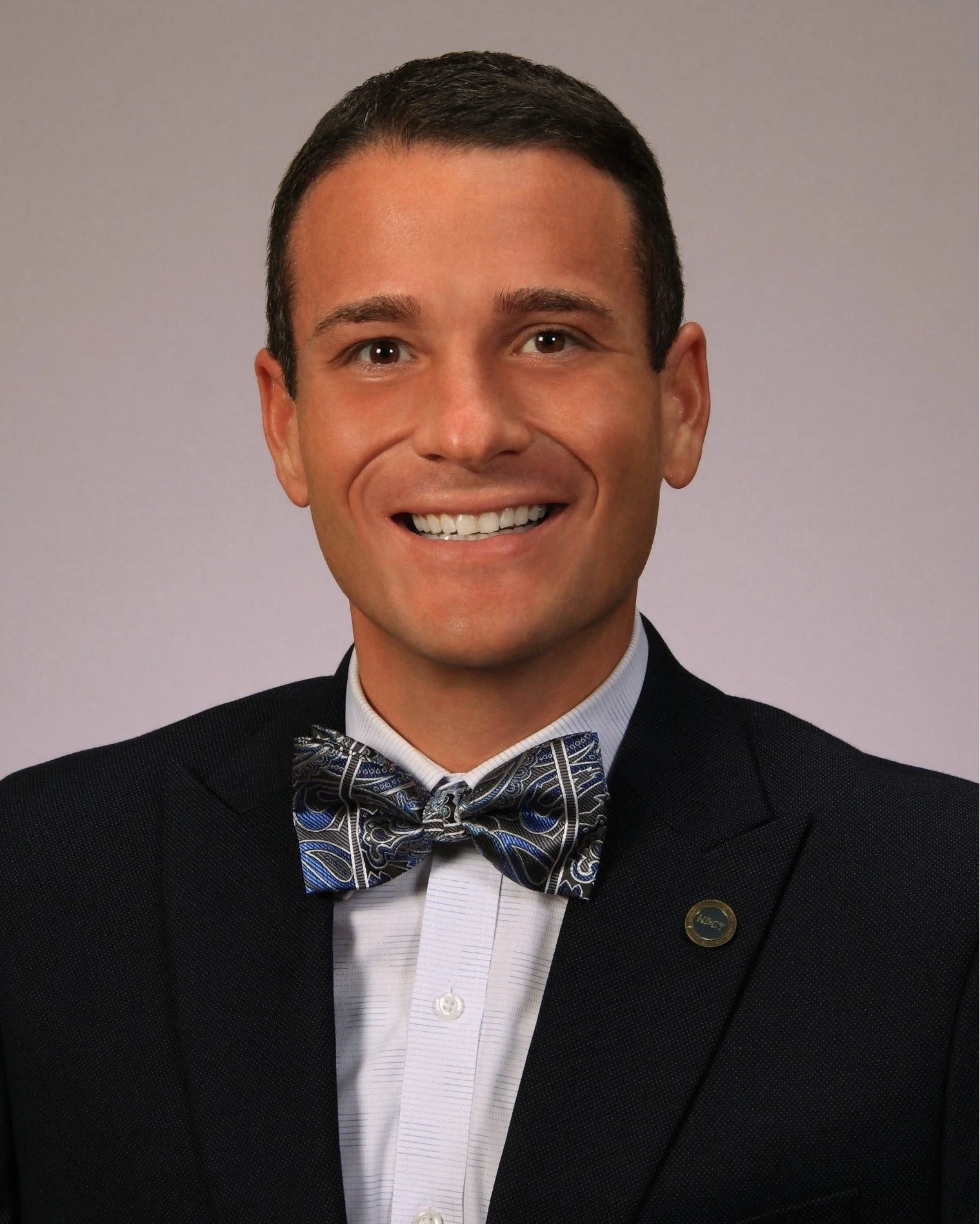 A smiling man in a dark suit and patterned bow tie poses for a professional headshot against a light background.