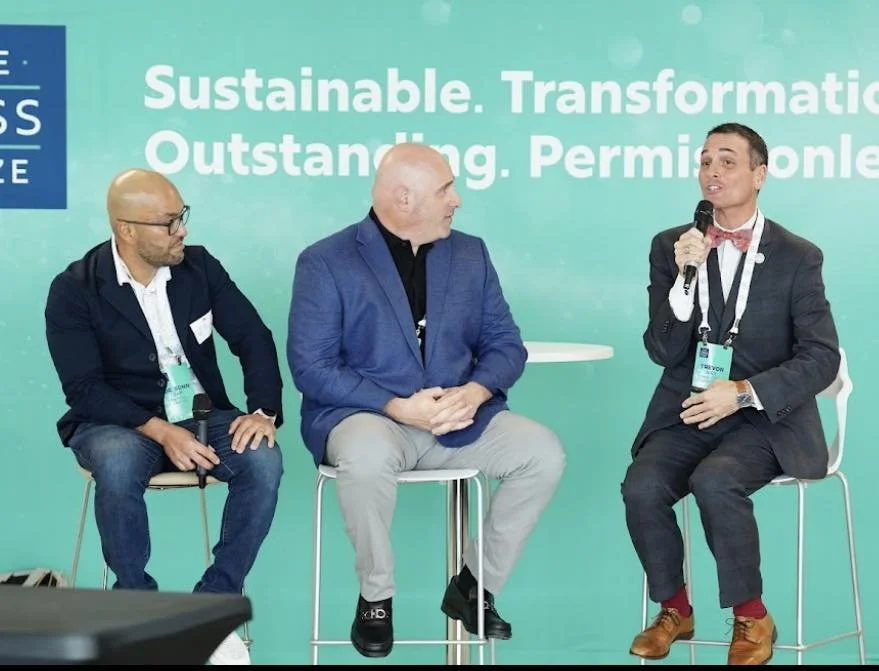 Three panelists sit on stools in front of a teal conference backdrop while one of them speaks into a microphone, wearing a suit and bow tie with a conference badge.