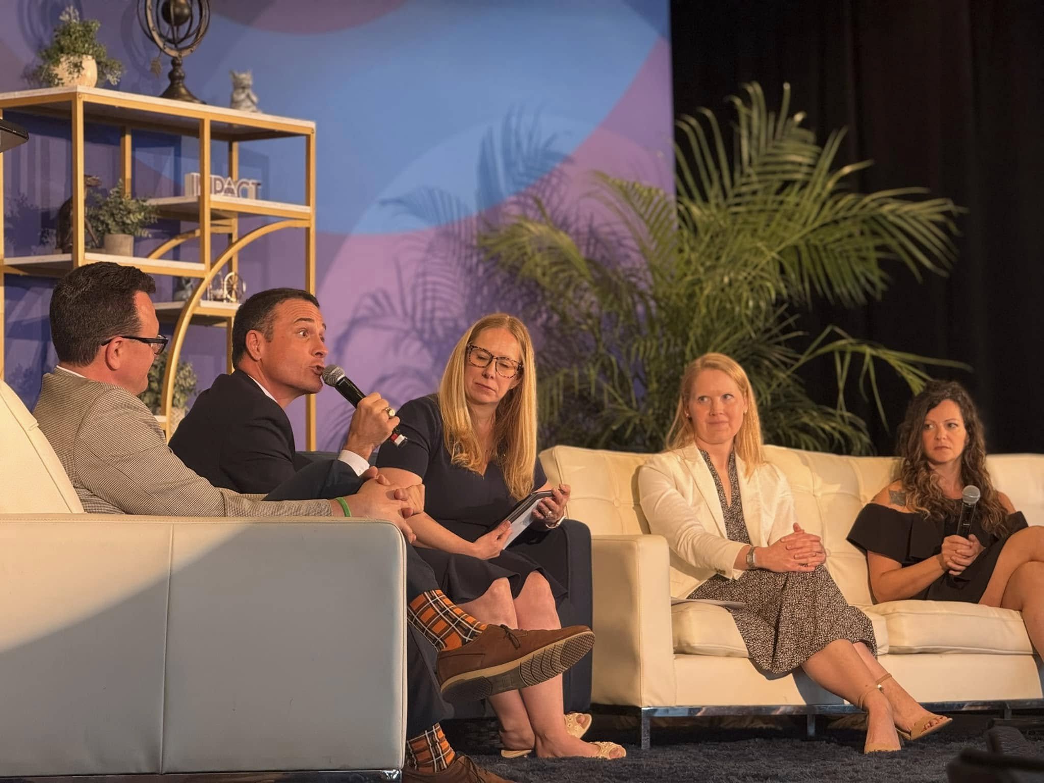 A panel of five people sit on stage having a discussion, with one man holding a microphone while speaking. They are seated on white couches with decorative shelving and plants in the background.