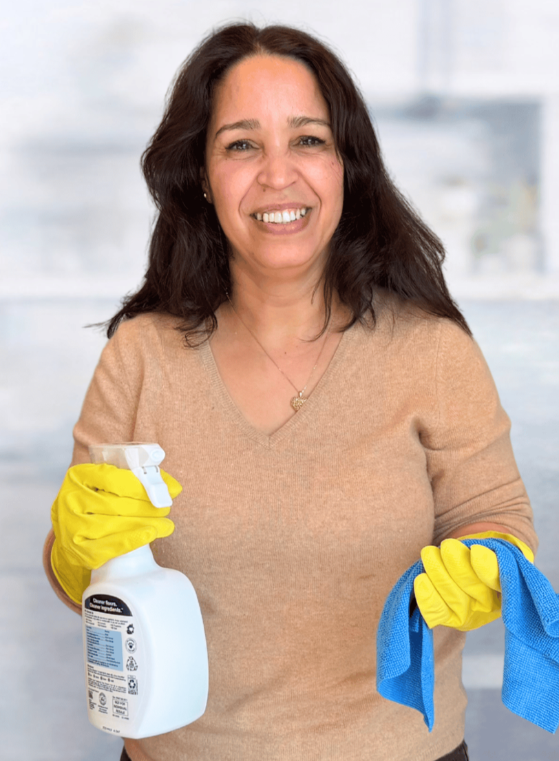 A woman smiling, wearing a beige sweater, yellow rubber gloves, and holding a spray bottle of cleaning solution in one hand and a blue cleaning cloth in the other, in a bright room.