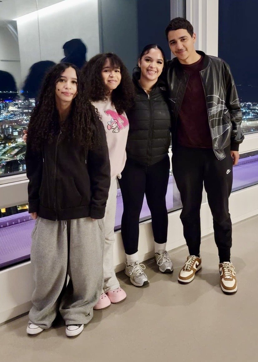 Four young people standing together indoors at night, with a city skyline visible through the window behind them. The group includes one young woman with long curly hair, another woman with straight hair, and two young men, all smiling and wearing casual clothing.