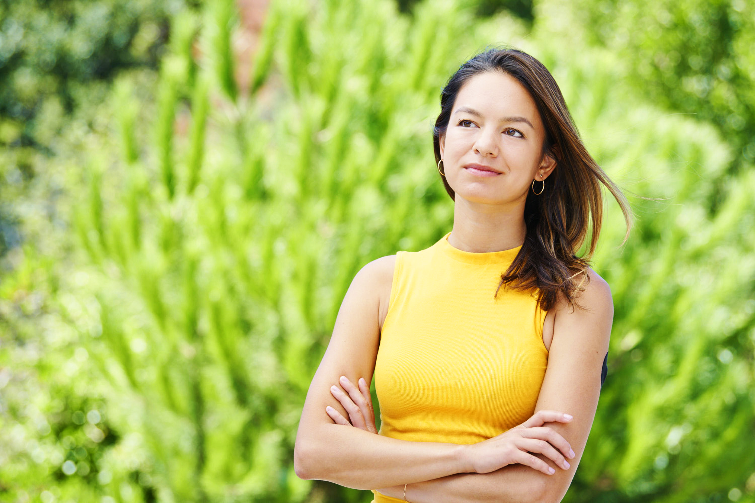 A woman in a yellow sleeveless top standing outdoors with crossed arms, smiling and looking to the side, with green foliage in the background.