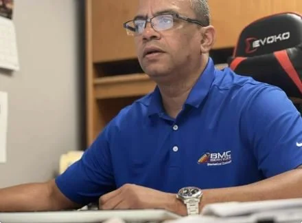 Man wearing glasses and a blue polo shirt, sitting at a desk with his arm on the surface, in a room with a chair and wooden shelves behind him
