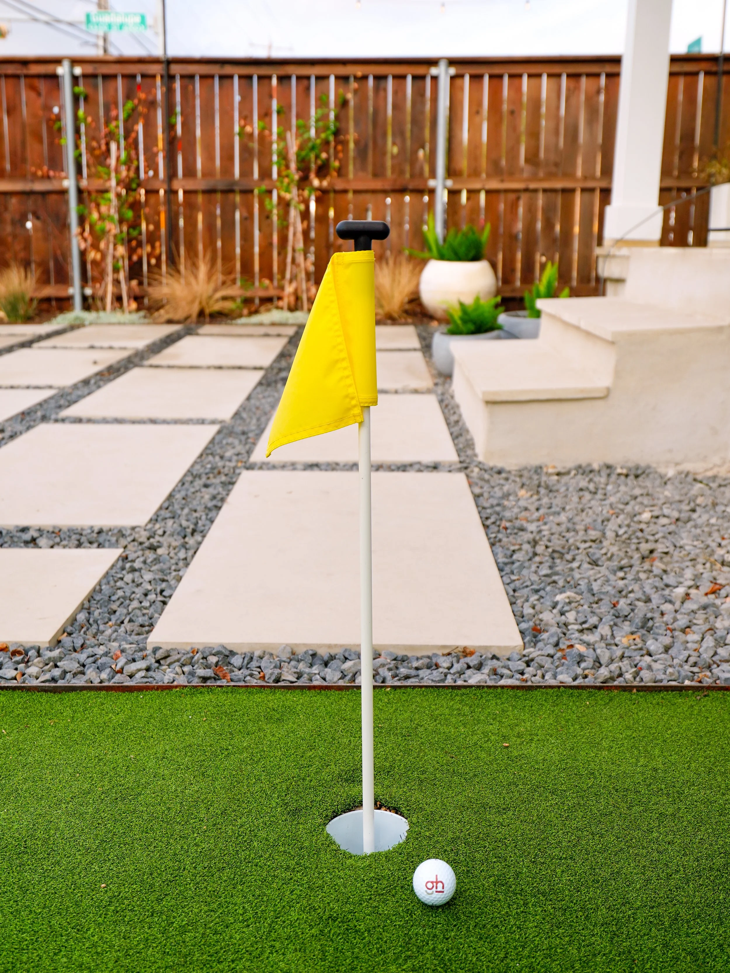 Miniature golf putting green with a ball, flag, and hole, in a backyard with a wooden fence, potted plants, and concrete steps.