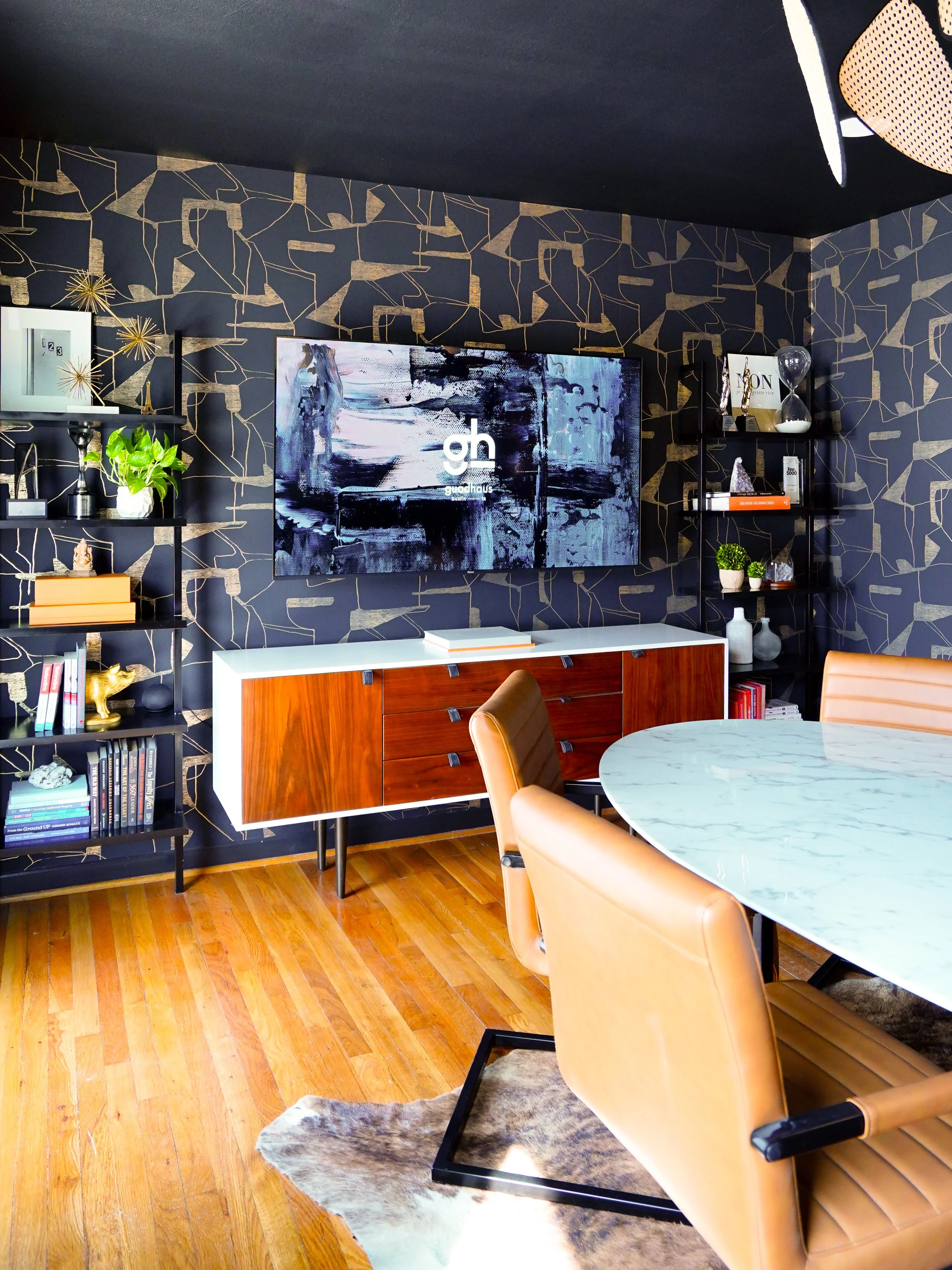 Modern living room with a black and gold abstract patterned accent wall, a mounted TV, black shelving units with decor, a wooden sideboard, tan leather chairs, a marble tabletop, and hardwood floors.