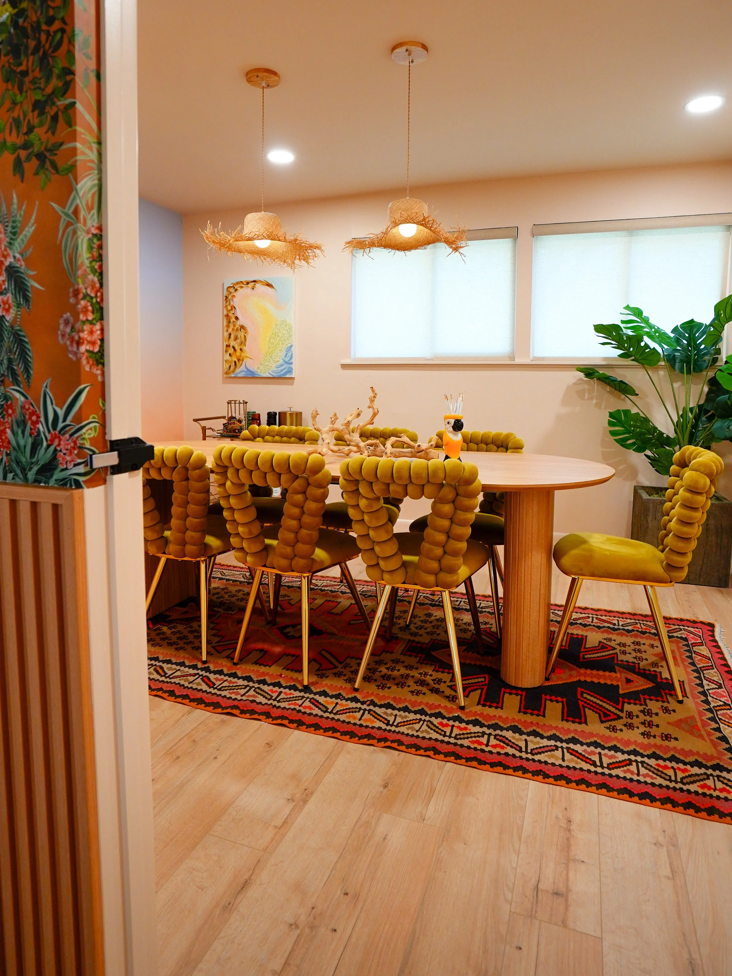 A dining room with a wooden table and six yellow padded chairs with rounded cushions. Two hanging light fixtures with woven shades are above the table. There is a colorful rug underneath the table, a large green plant in a pot on the right, and a window with white blinds in the background. A small decorative figurine and some objects are on the table.
