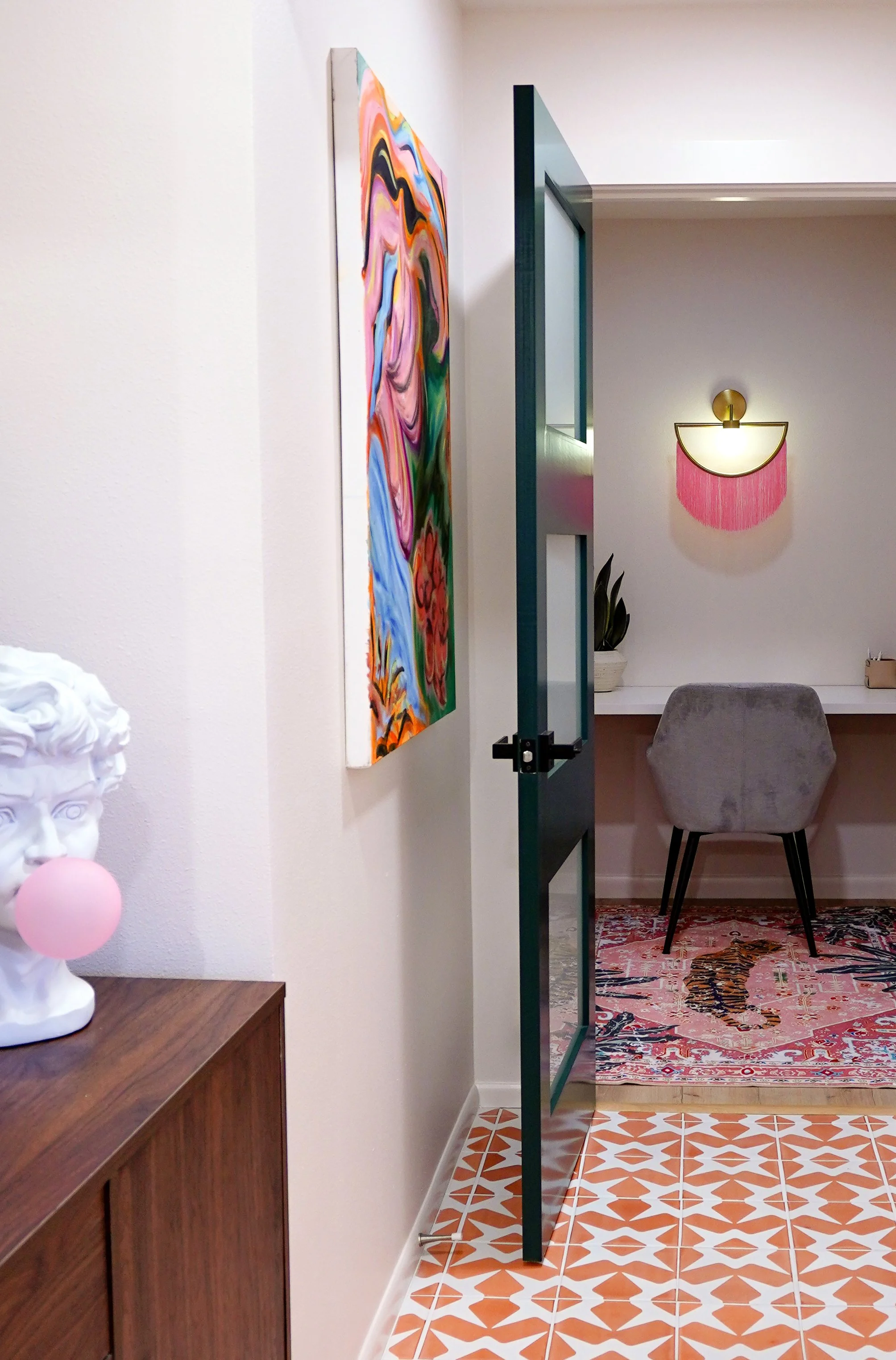 Interior view of a room with a colorful abstract painting on the wall, a small desk with a chair, a wall sconce with pink fringe, and a patterned rug featuring a tiger design.