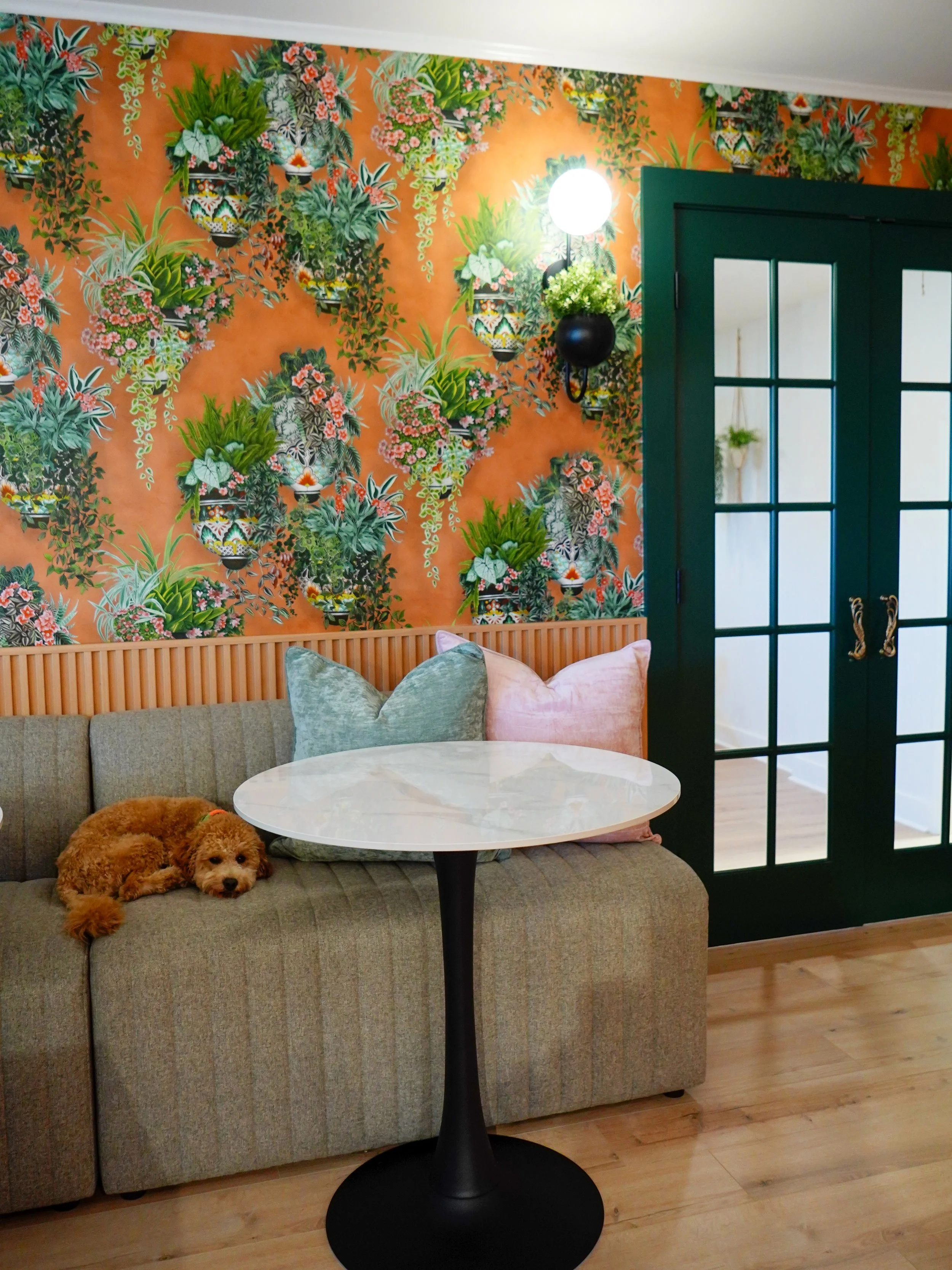 A cozy living room with a green couch featuring pink and gray pillows and a small brown dog lying on it. Behind the couch is a vibrant, floral wallpaper with lush green plants, pink flowers, and decorative pots. To the right, there are glass-paneled double doors painted dark green, with a white wall visible through the glass. A round white table with a black pedestal base is in front of the couch.