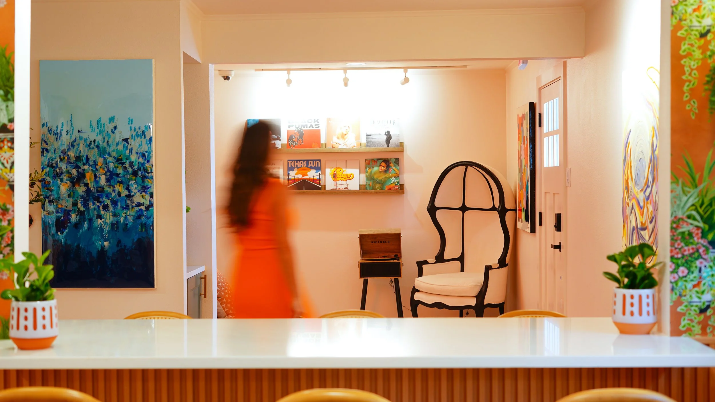 A cozy interior with a woman in an orange dress walking past a wall decorated with abstract artwork, a shelf with vinyl records, and a vintage armchair.