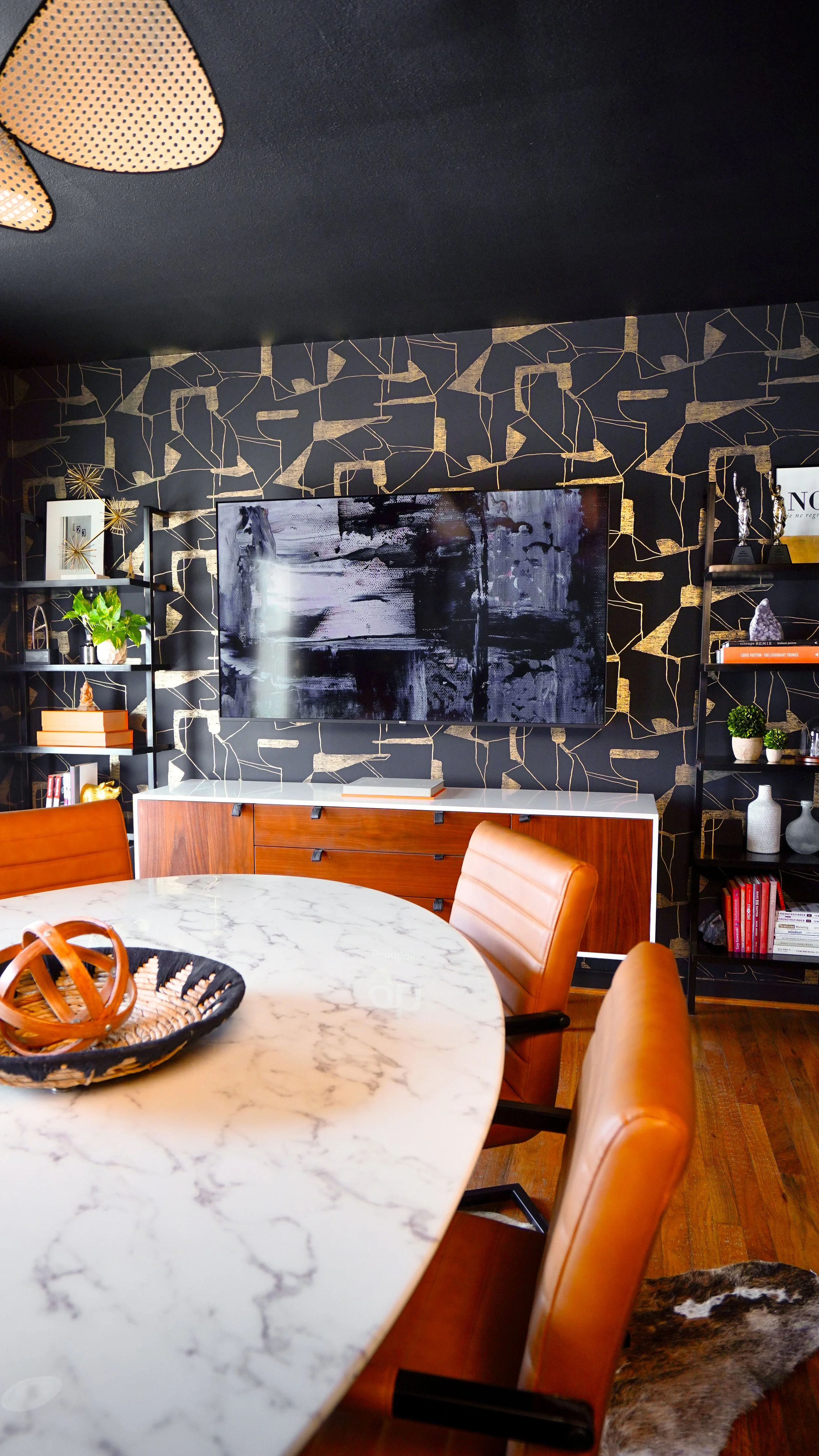 Modern dining room with a marble table, tan leather chairs, black bookshelves with decorative objects, and a wall-mounted television on a black and gold patterned wall.