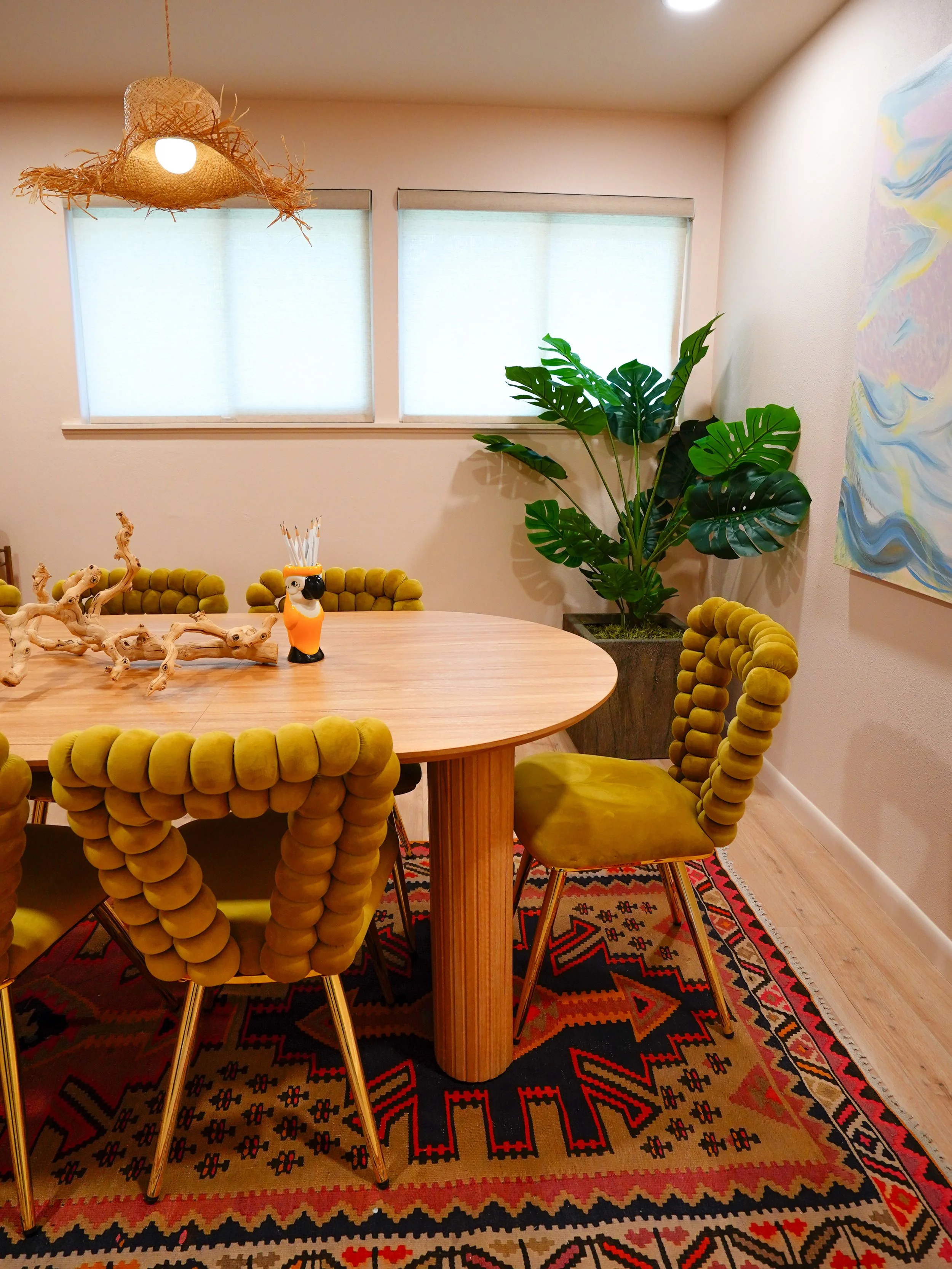 A dining room with a wooden table and six green upholstered chairs with unique rounded armrests. There is a colorful patterned rug under the table, a large green potted plant in the corner, and abstract art on the wall. Two windows with light-colored