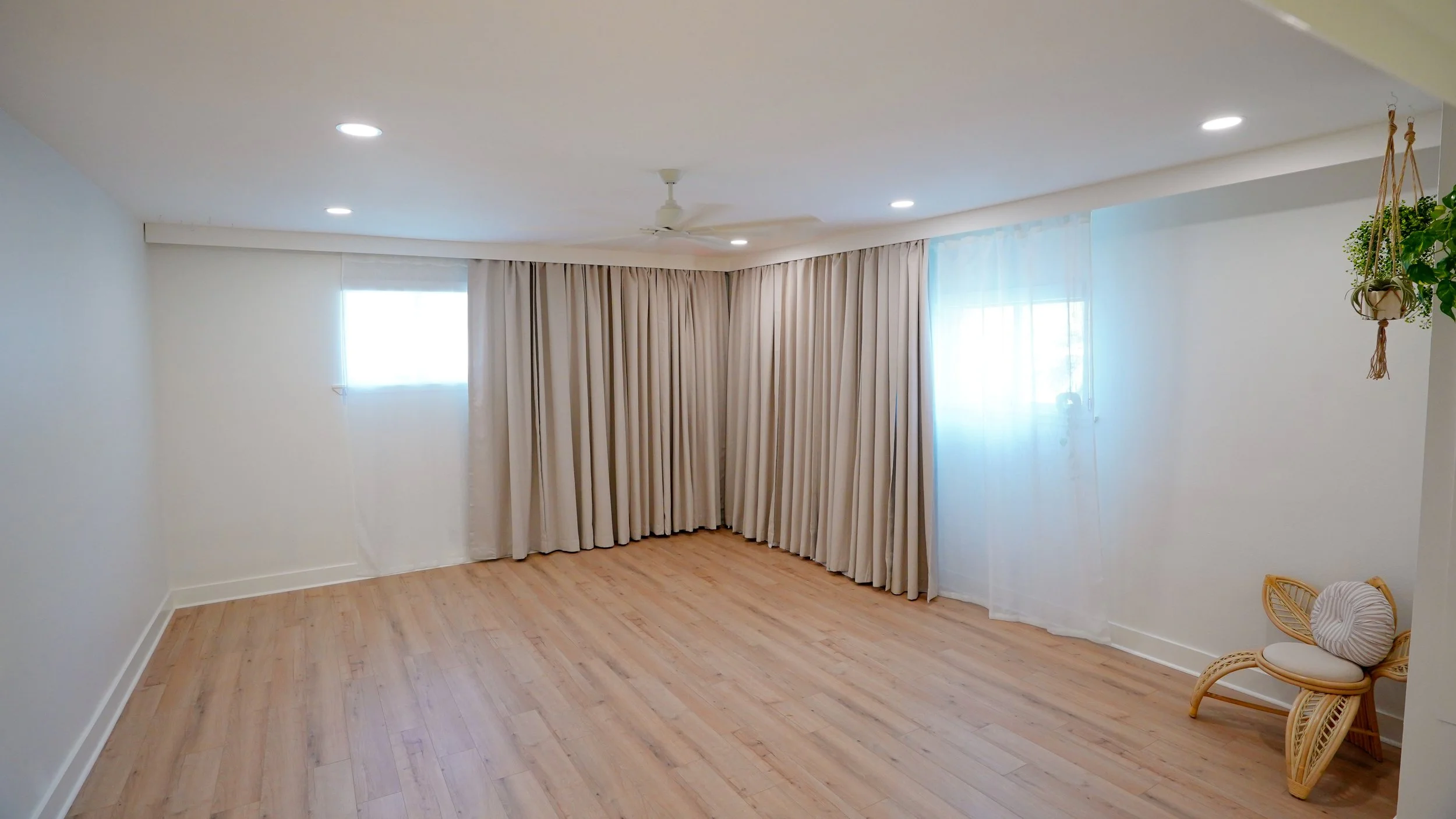 Empty room with beige curtains, sheer curtains, hardwood floors, ceiling fans, and a wicker chair with pillow in the corner.