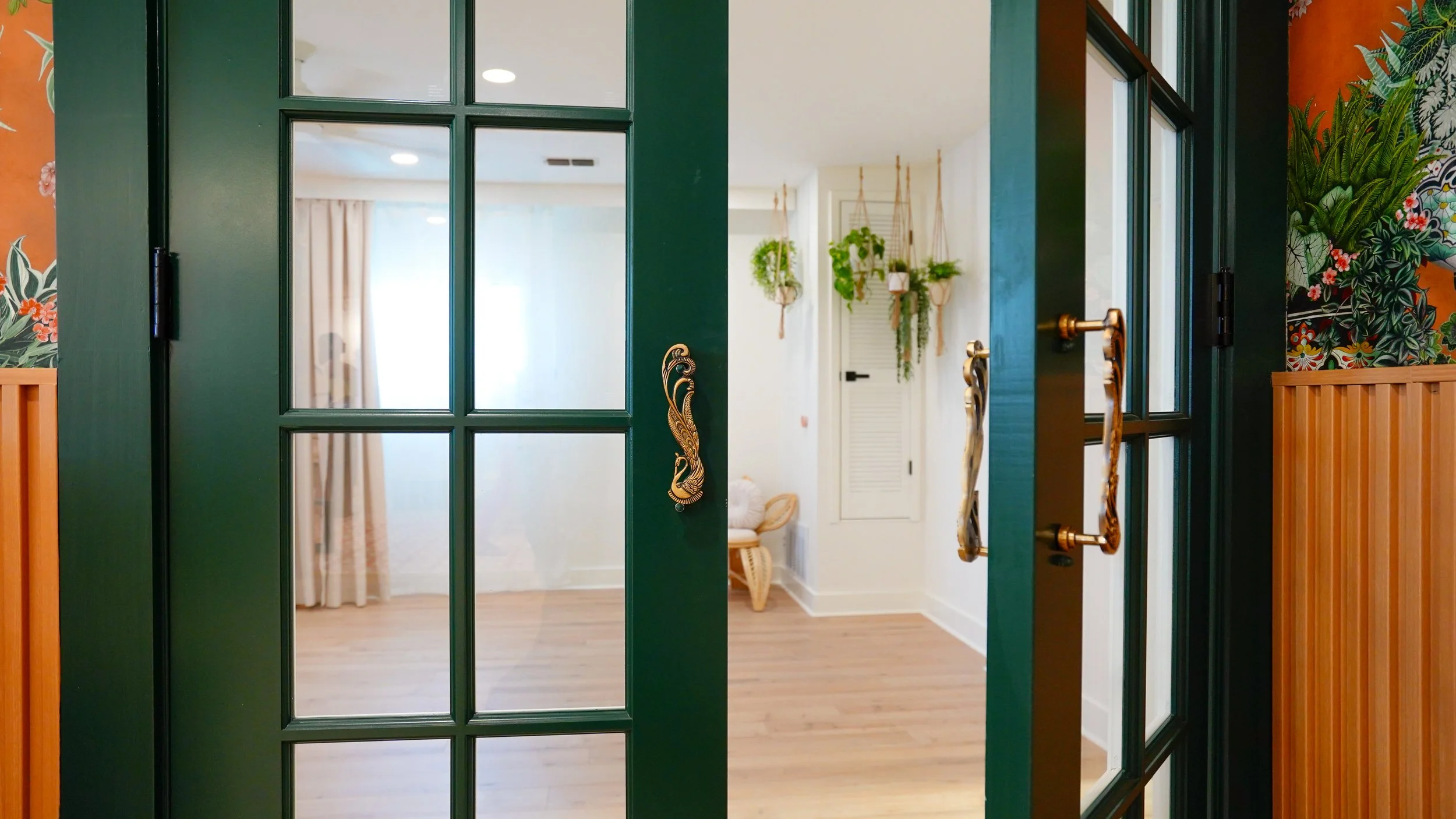 Open green French doors with gold handles leading into a room with wooden flooring, hanging plants, and natural light.