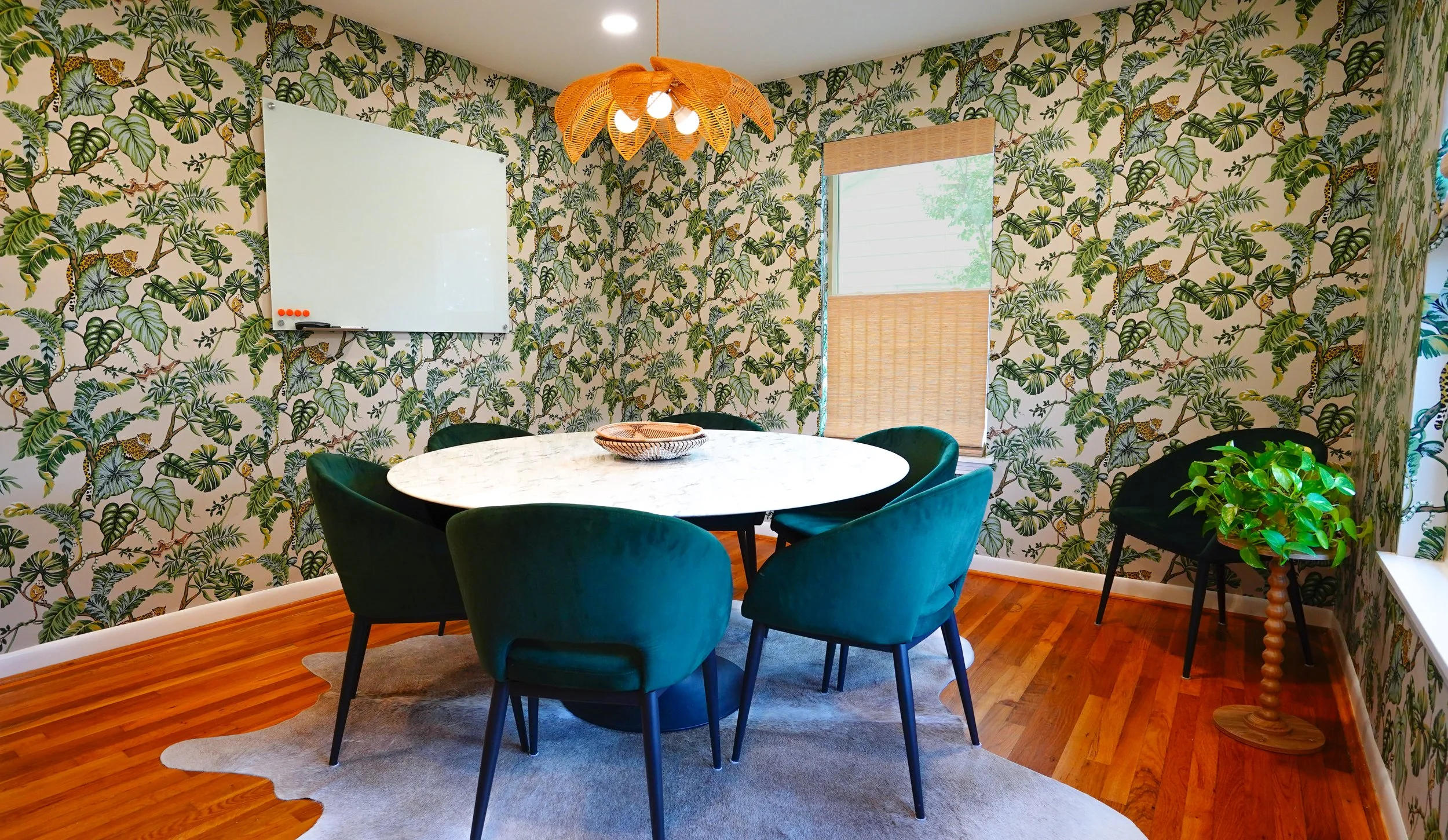 A dining room with jungle-themed wallpaper with leaves and animals, a round marble table with six green velvet chairs, a whiteboard on the wall, a window with blinds, a decorative pendant light, and a potted plant on a wooden stand.