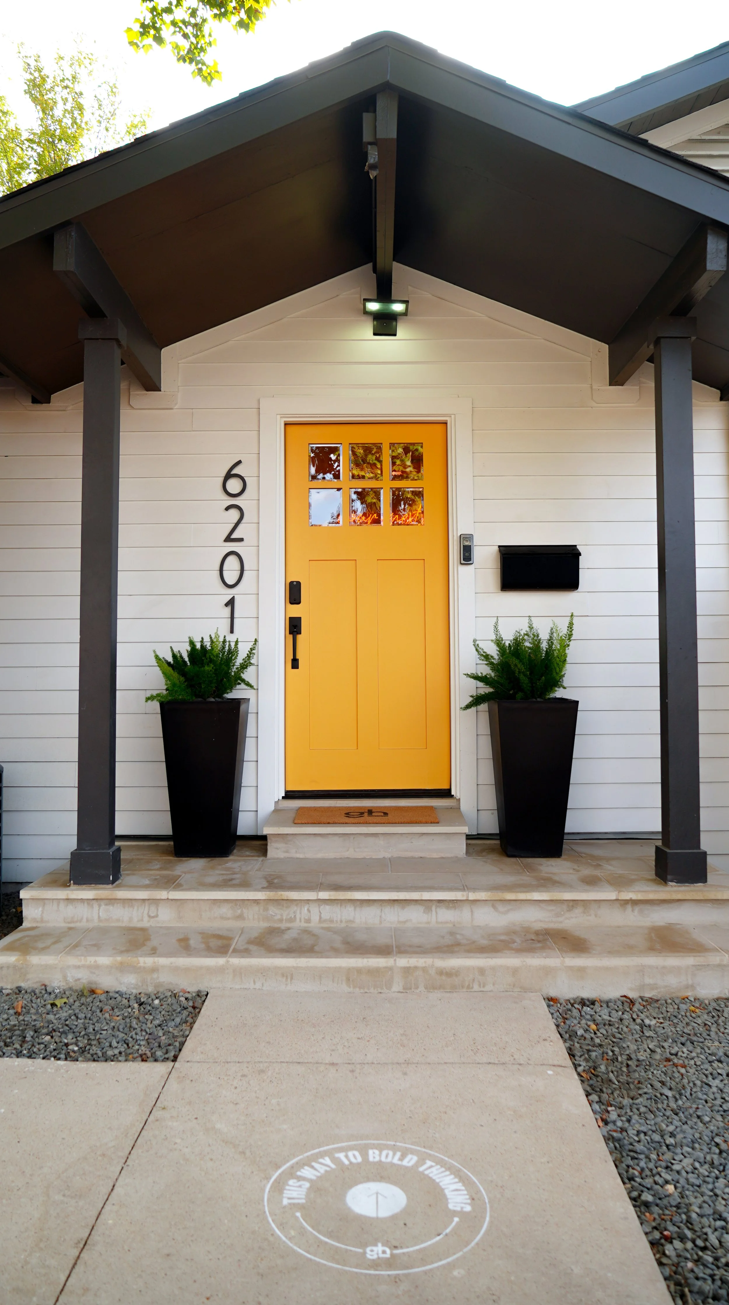 Front porch of a house with a yellow door, black mailbox, potted ferns, and house number 6201, under a black covered porch with a light.