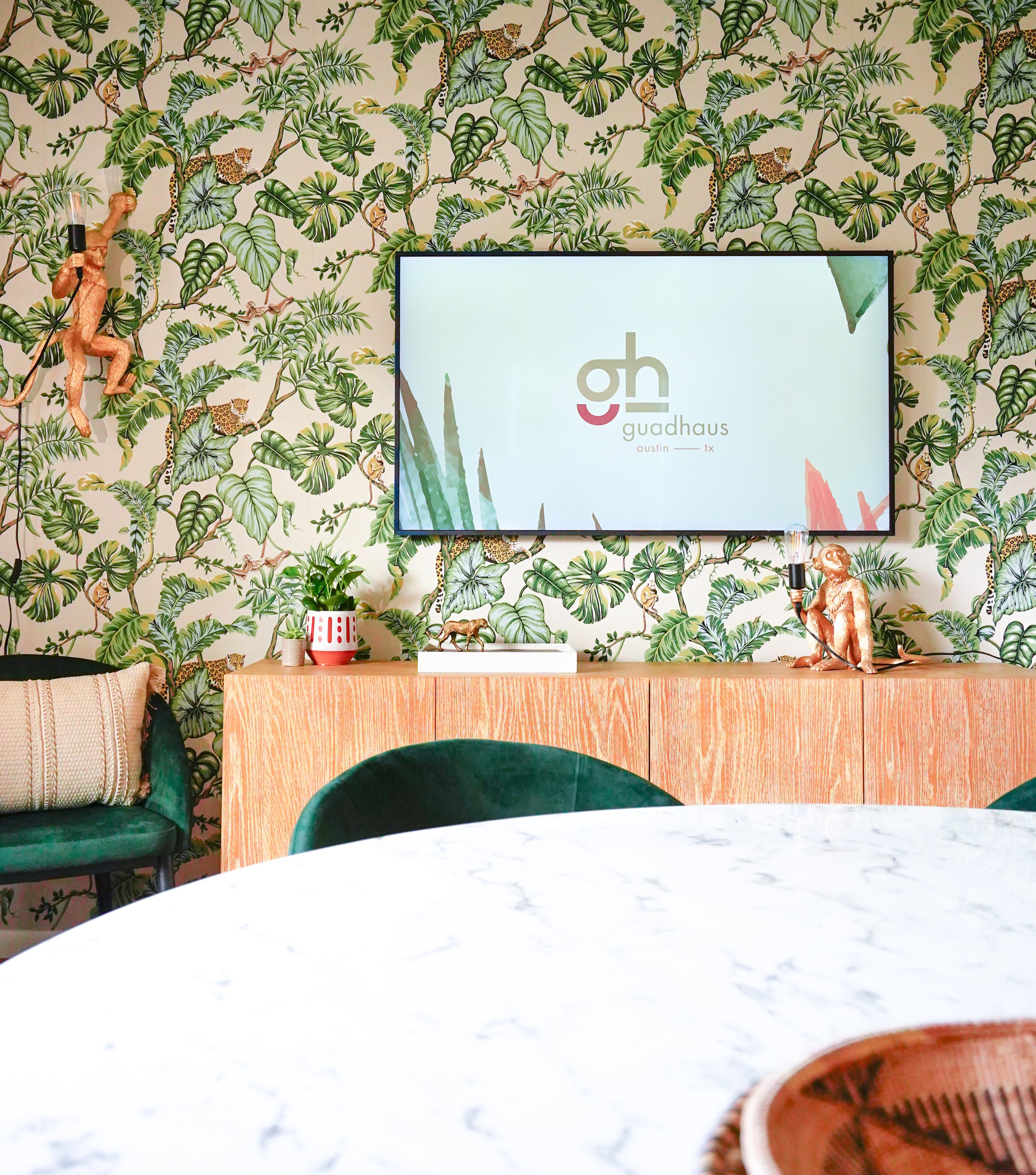 Interior room with tropical leaf and animal wallpaper, a television screen displaying 'guadhauxs,' a sideboard with plant and decorative items, and a marble-topped table in the foreground.