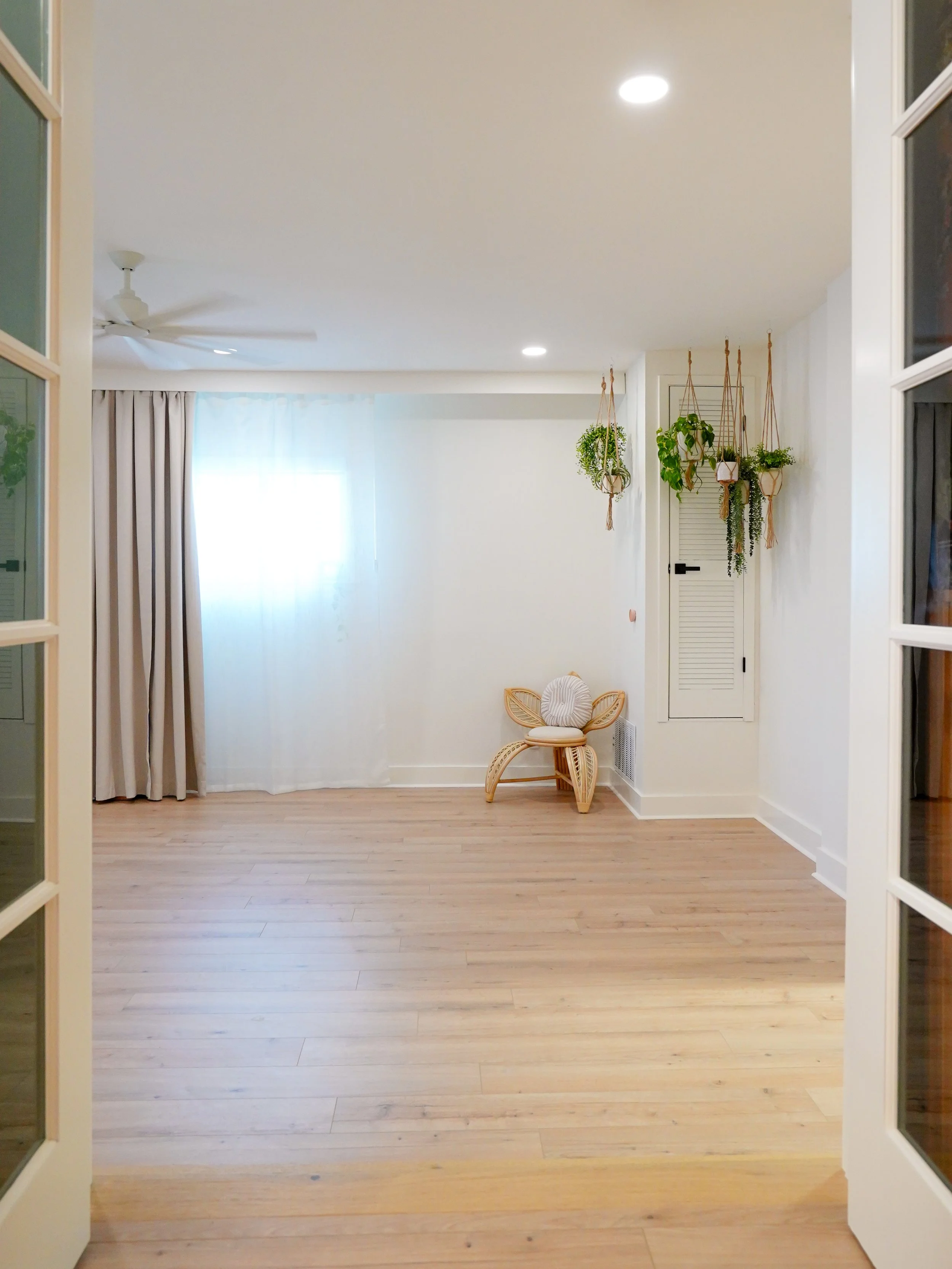 Empty room with wooden floor, white walls, hanging plants, beige curtains, a wicker chair with a cushion, closed closet door, ceiling fan, and recessed lighting.