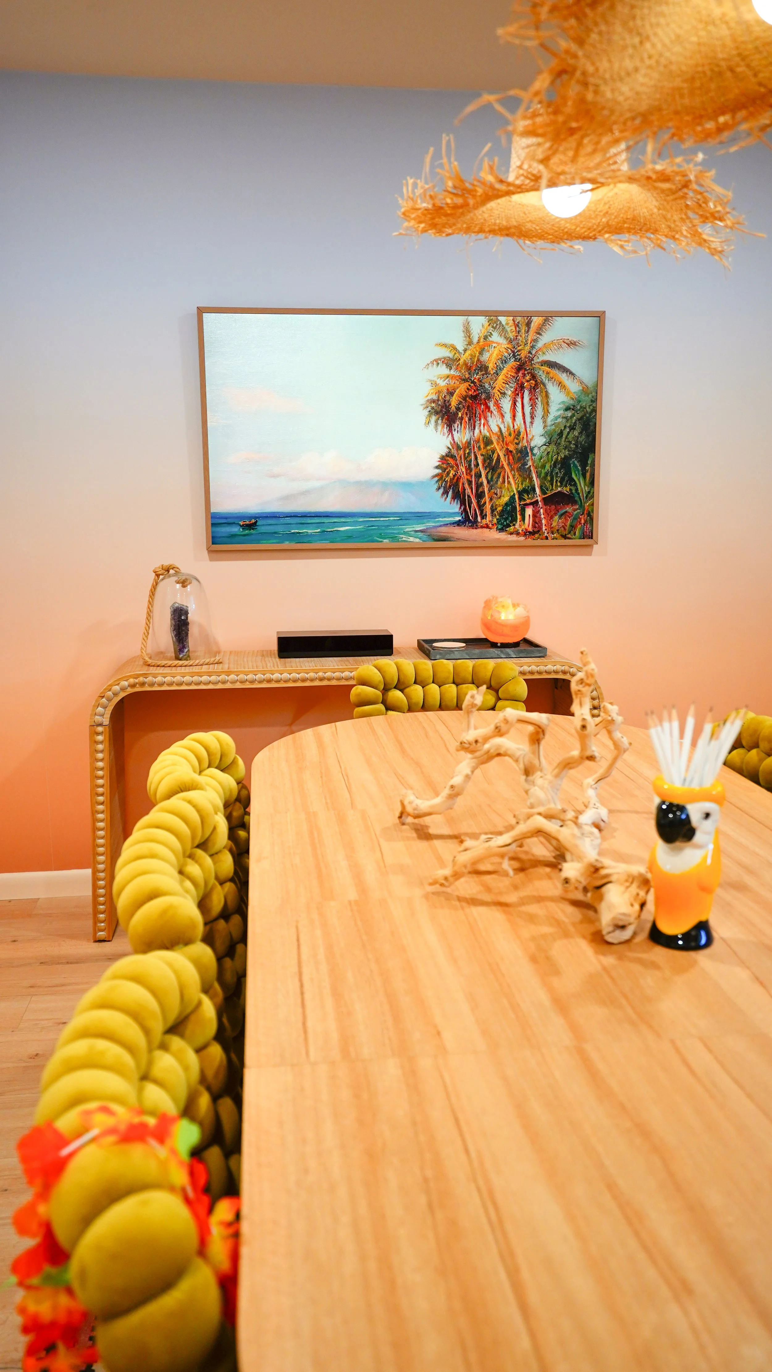 A dining room with a wooden table and yellow rounded chairs. On the table are decorative driftwood pieces and a dog-shaped container holding white straws. A buffet or sideboard with a glass jar and a salt lamp is against the wall. A framed painting o