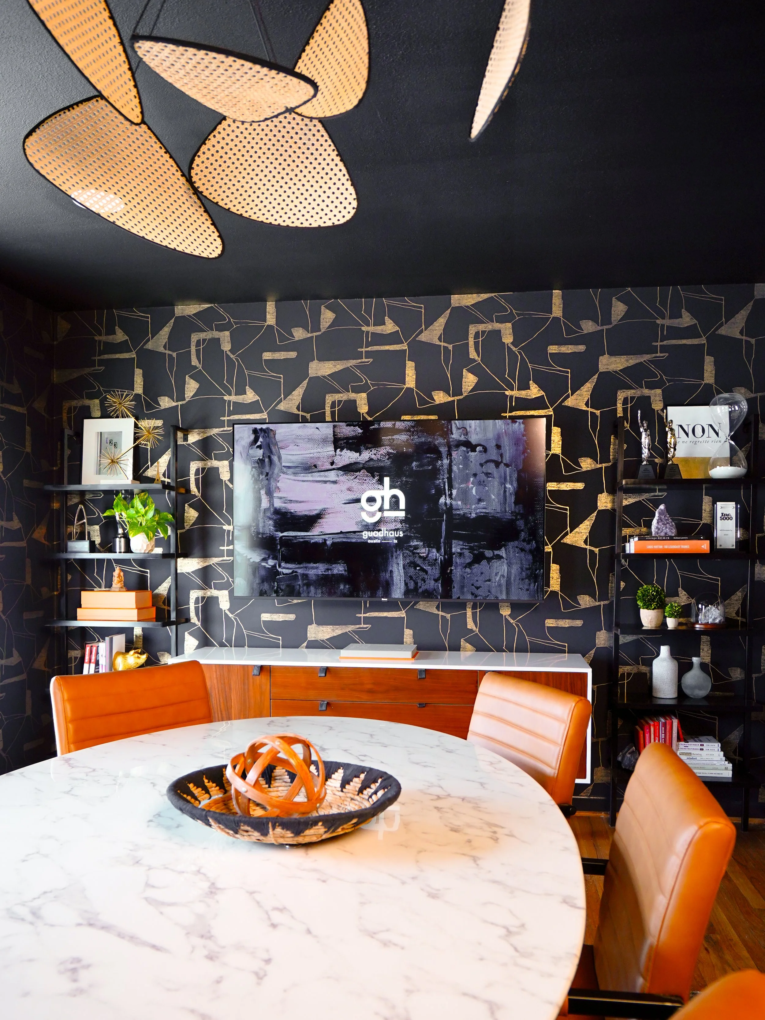 Modern dining room with a round marble table, leather chairs, and decorative shelving on a black wall with gold geometric patterns. A large TV displays the logo 'guudhaus'.