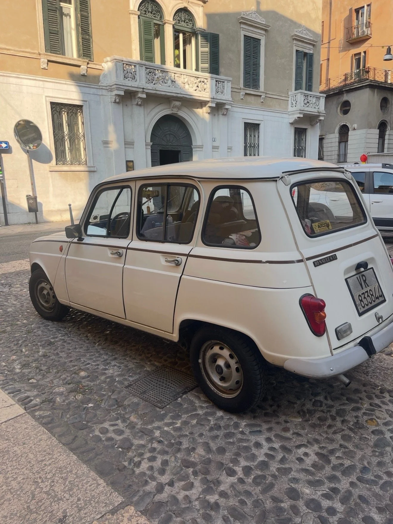 Vintage white Renault 4TL car parked on a cobblestone street in front of historic European-style buildings with green shutters.