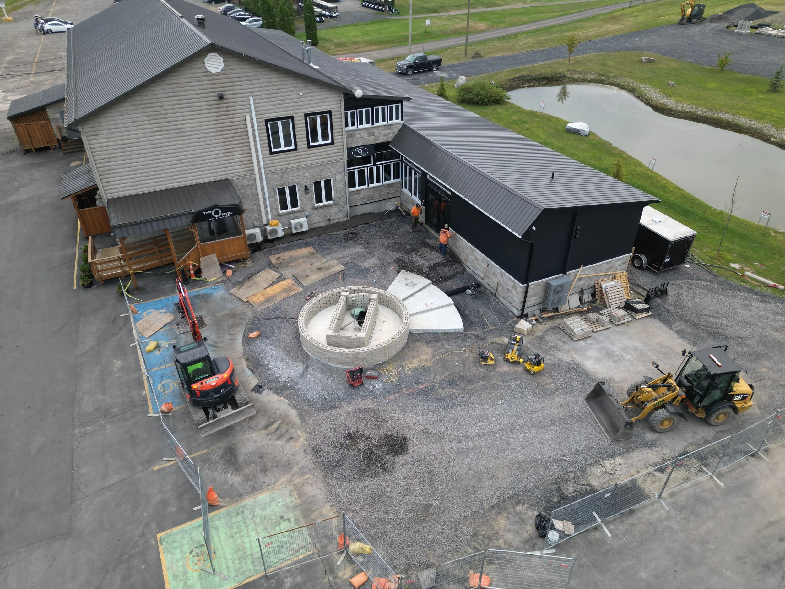 Construction site for a building expansion or renovation with construction workers, machinery, and materials, near a pond and parking lot.