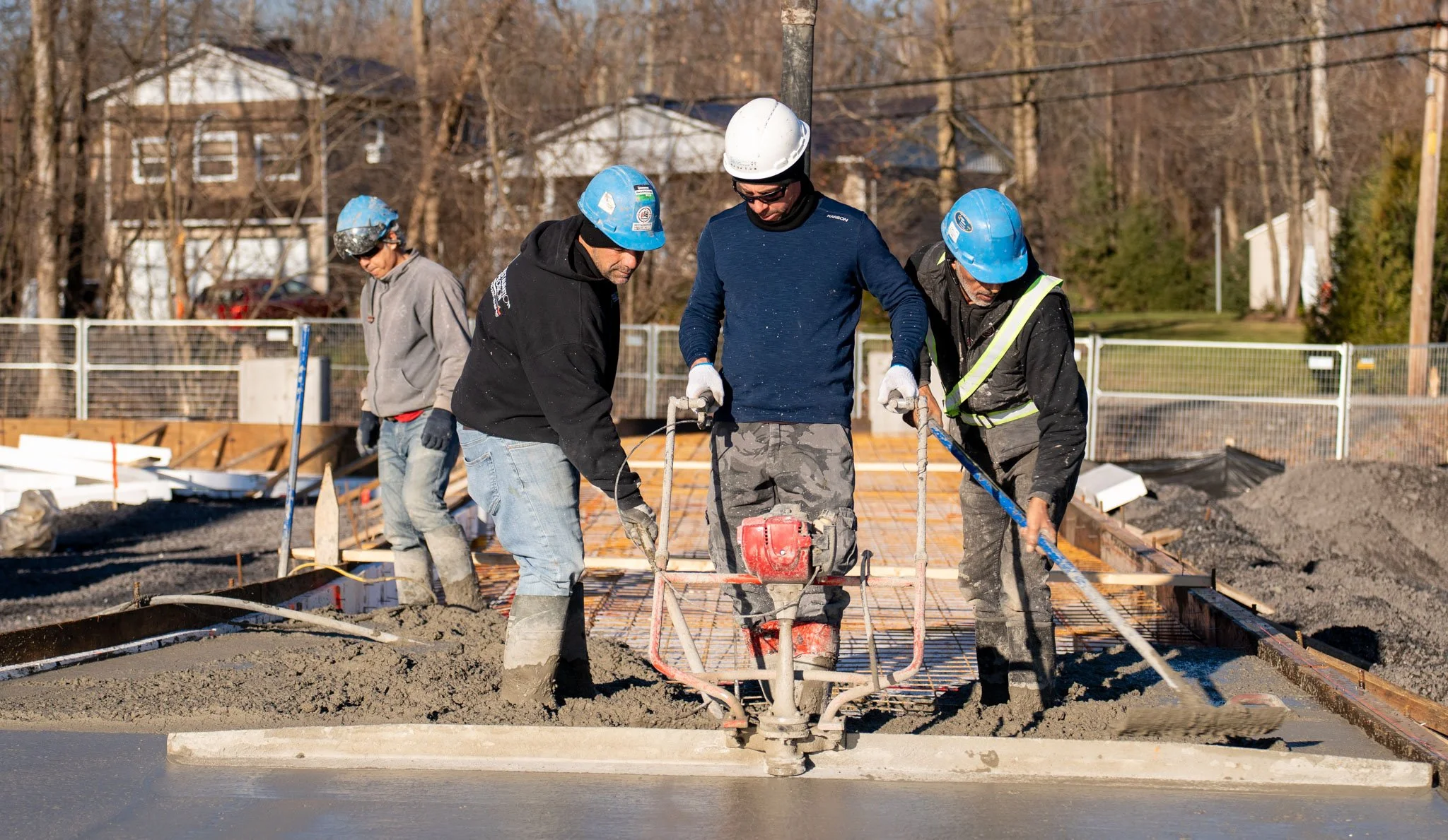 Construction workers in safety helmets and gloves working on pouring concrete on a building site with houses and trees in the background.