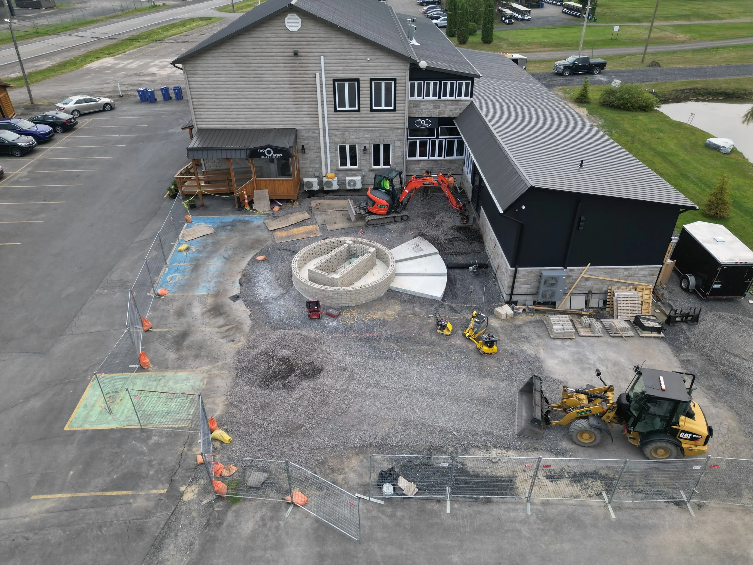 Construction site with a building, construction equipment, and materials. Fencing surrounds the area, with paving work in progress on the ground. Vehicles are parked nearby, and there is a pond in the background.
