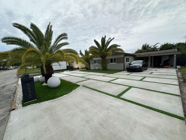 Modern house with a concrete driveway divided by grass strips, palm trees, and a black car parked in the driveway.