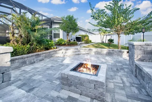 Outdoor patio with fire pit, stone walls, lush green trees, plants, and a house in the background under a partly cloudy sky.