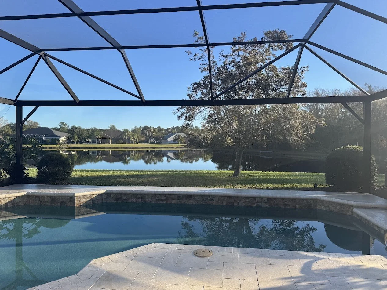 View of backyard with pool, lake, trees, and houses in the distance, under a screened enclosure.