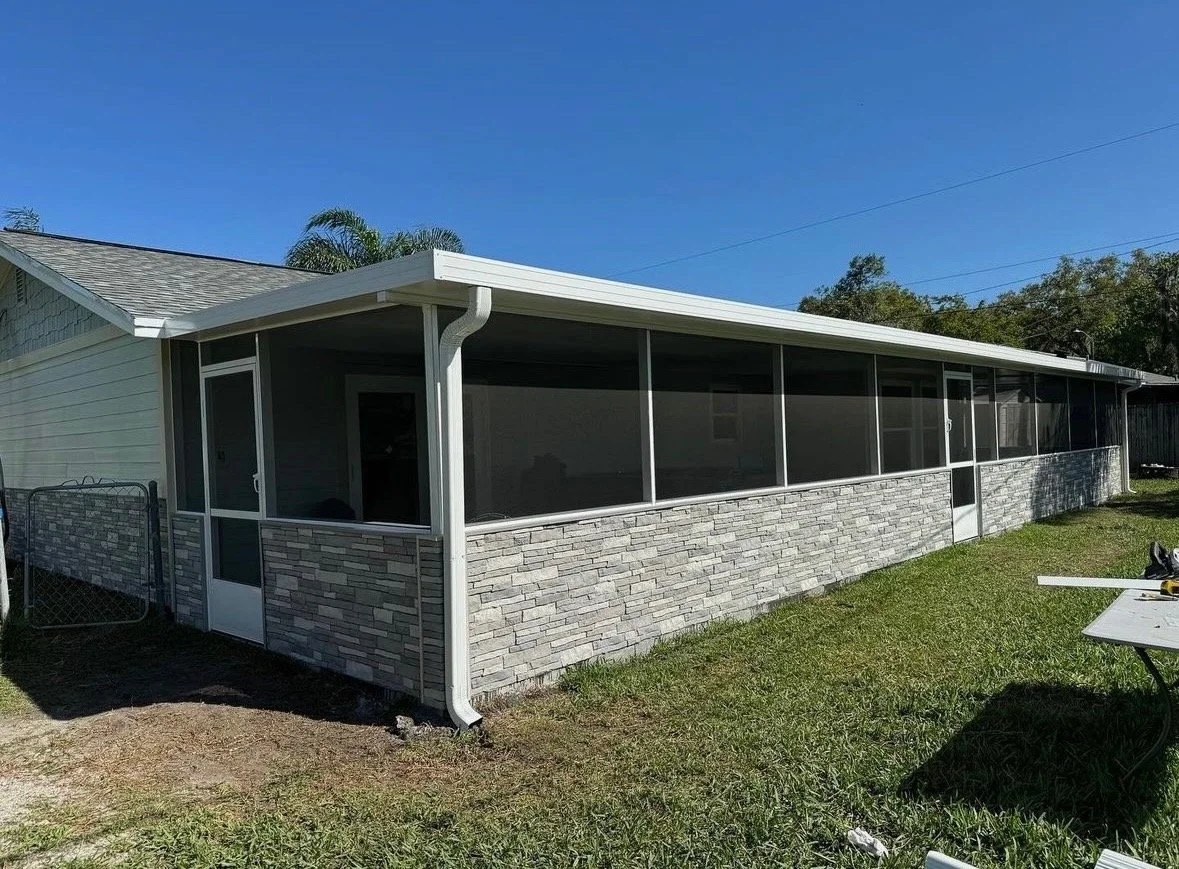 A backyard screened porch with stone veneer on the lower half, white framing, a white door, and a gray roof, adjacent to a house under clear blue sky.