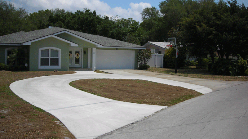 Newly paved winding driveway leading to a single-story house with a two-car garage, green lawn, surrounded by trees.