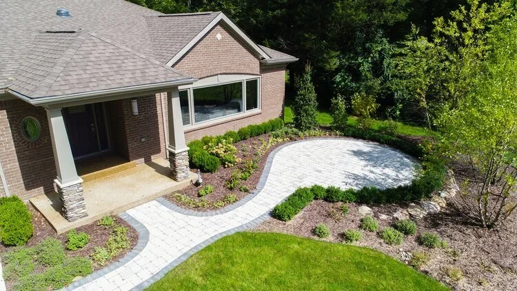 A front yard with a brick house, a paved walkway, a small lawn, and landscaped garden beds with bushes and trees.