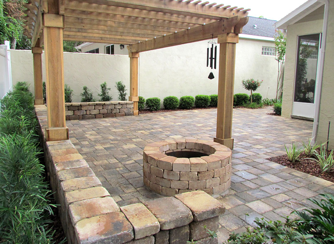 Backyard patio with a stone fire pit, brick border, wooden pergola, wind chimes, and various plants.