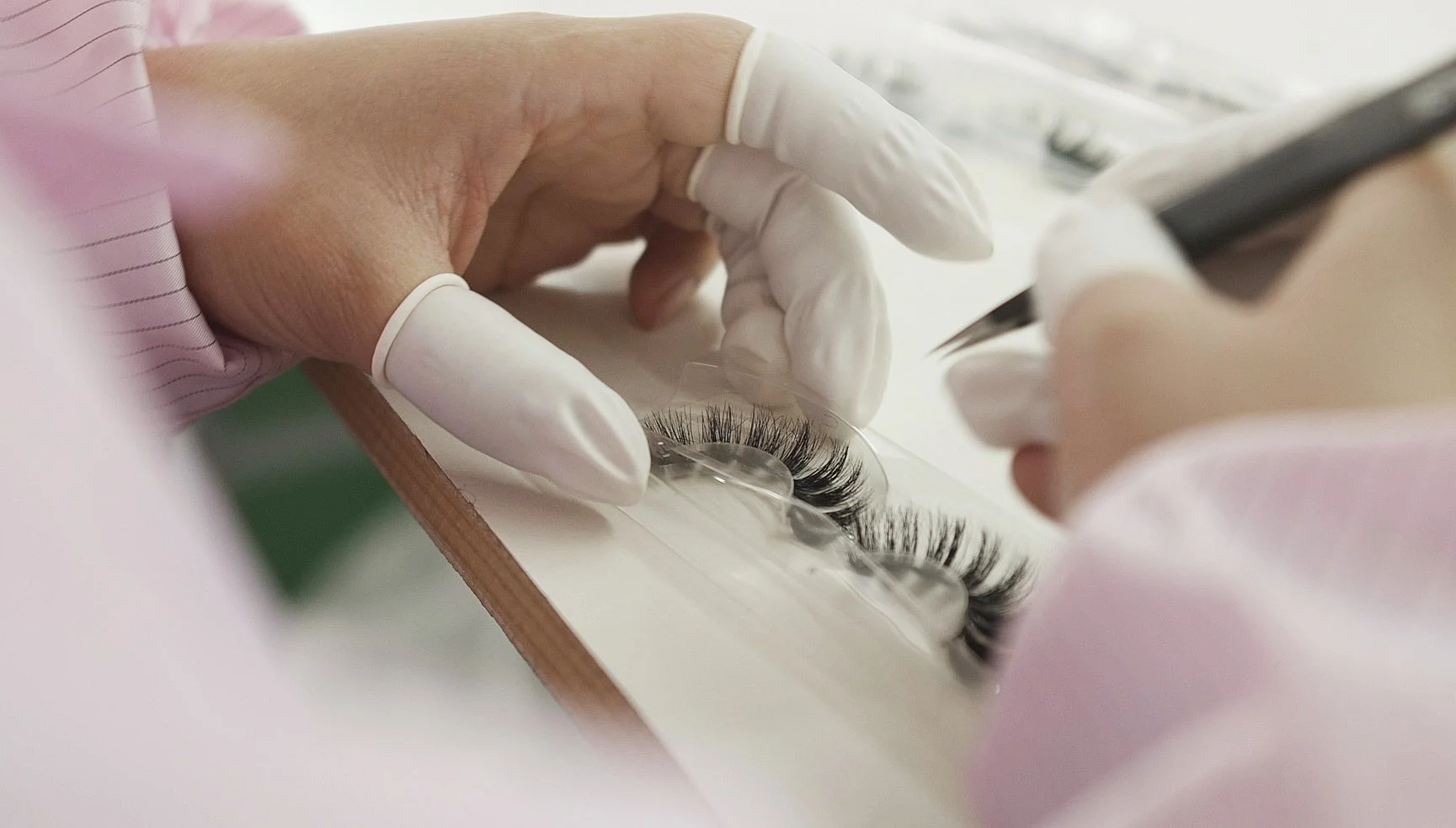 Close-up of a person's hands wearing white gloves attaching false eyelashes to a clear plastic eyelash strip at a workspace.