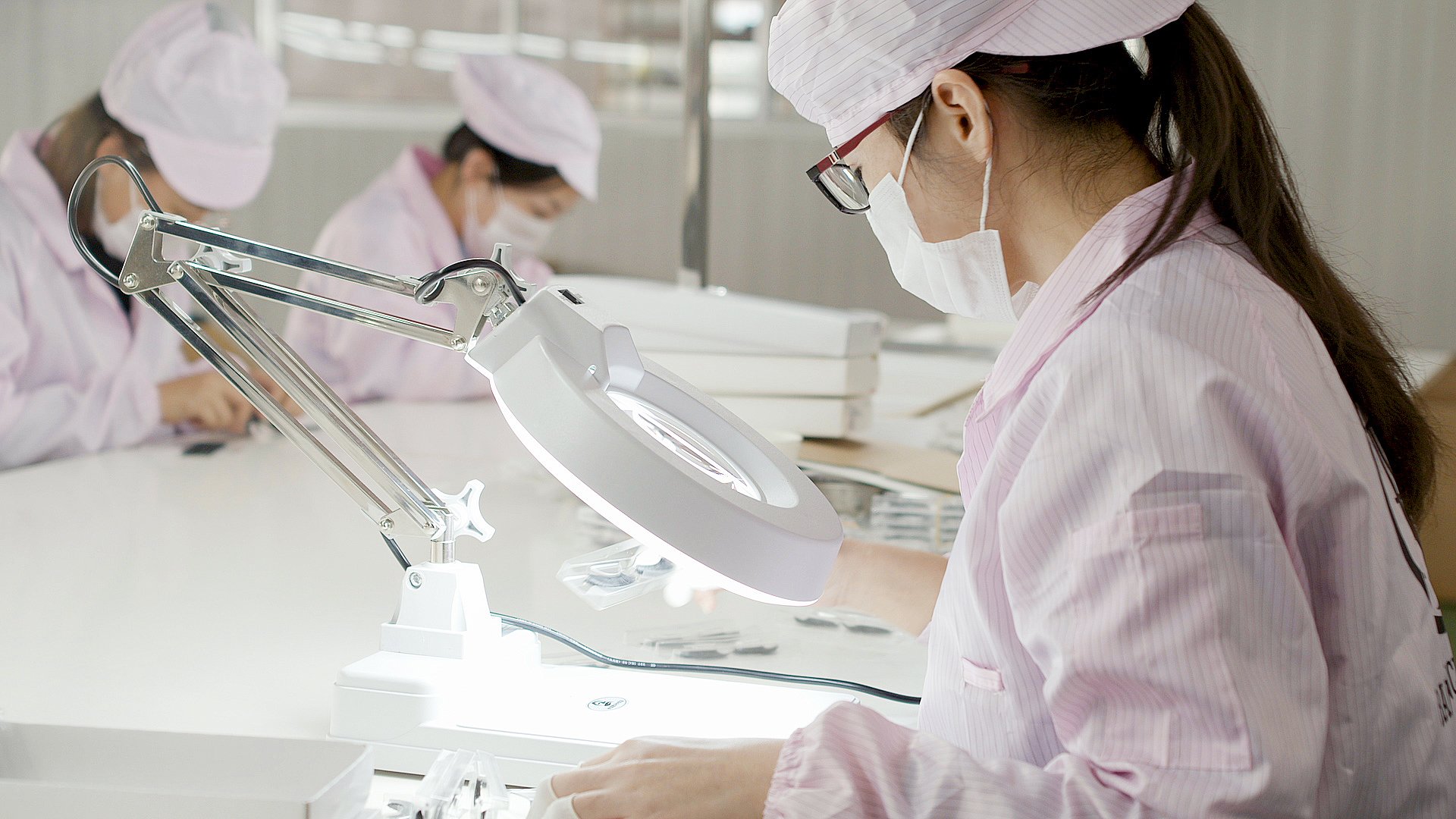 Female healthcare workers or scientists in pink uniforms, masks, and glasses working at laboratory stations with magnifying lamps.