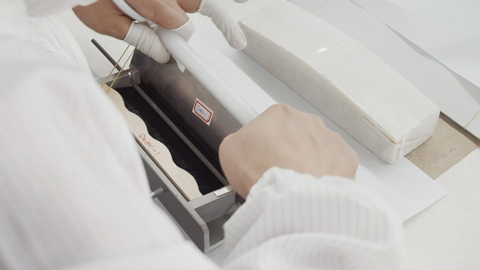 A person wearing white gloves using a machine to work on a material, possibly a lab or industrial setting, with a rectangular foam block and a wooden piece nearby.