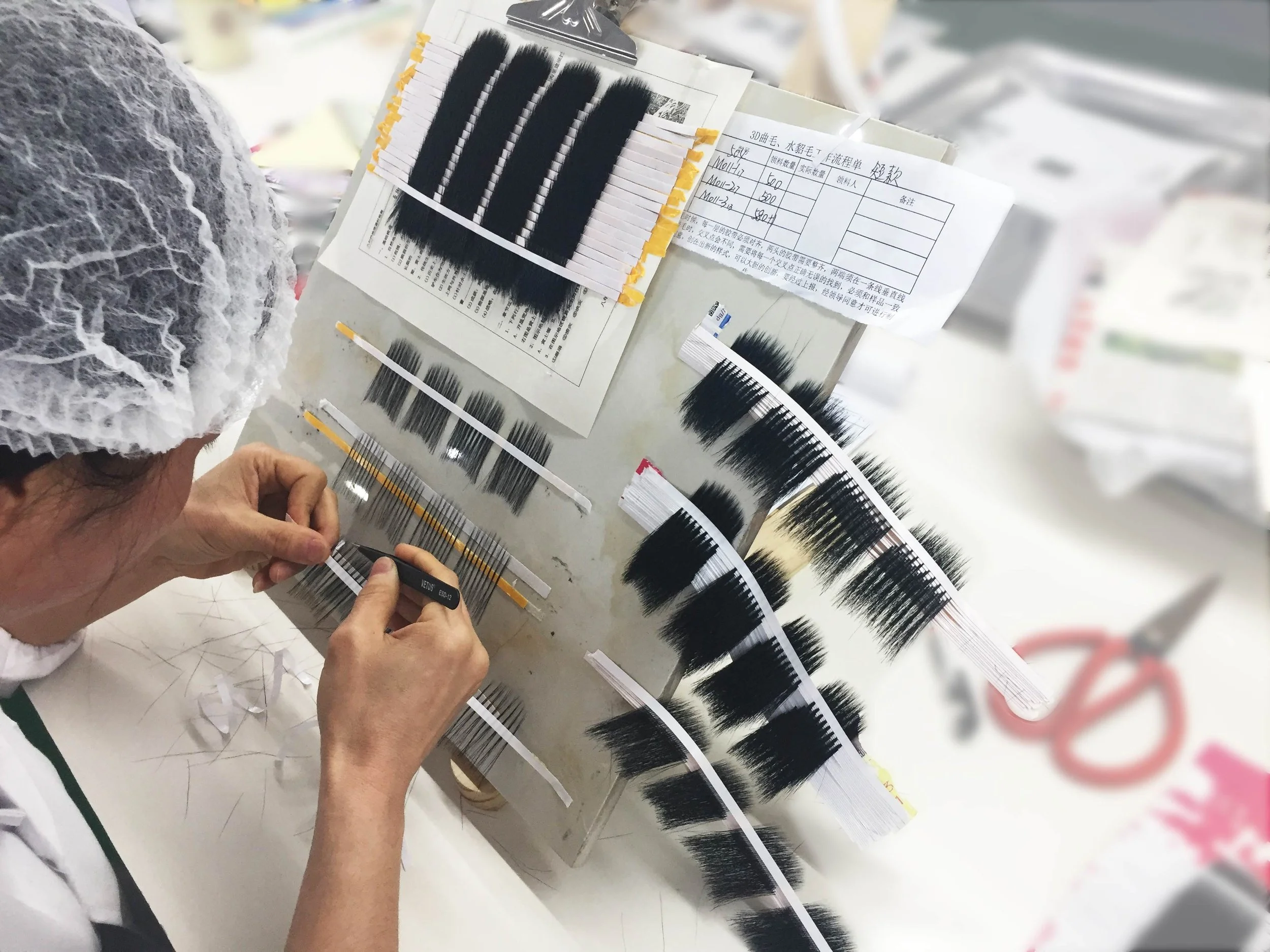 Person inspecting and handling false eyelash samples in a manufacturing or testing facility, with several rows of false eyelash strips and testing data sheets on the table.